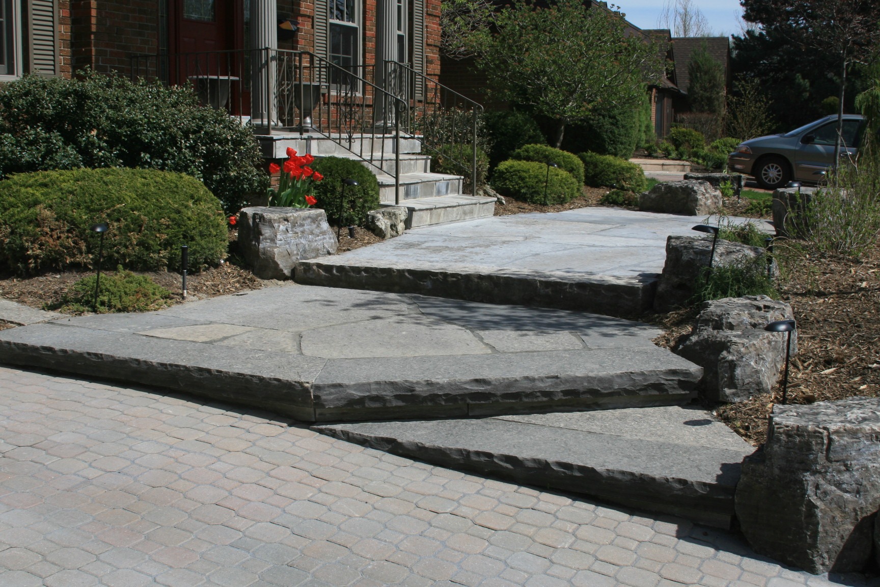 Stone steps and walkway lead to a brick house entrance, surrounded by greenery and a parked car, on a sunny day.