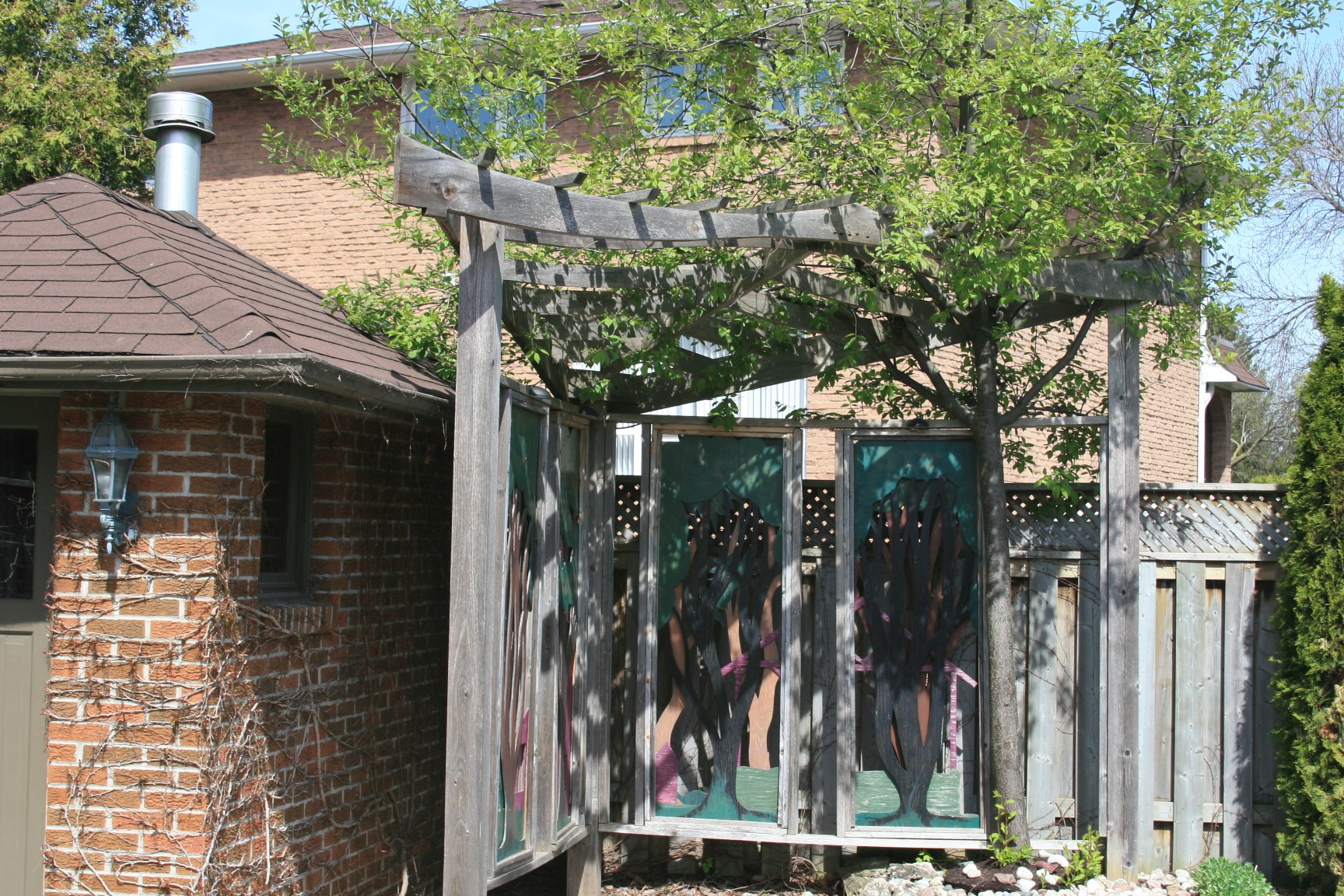 A garden scene features a wooden trellis with stained glass panels, beside a brick house and wooden fence, under a lush tree.