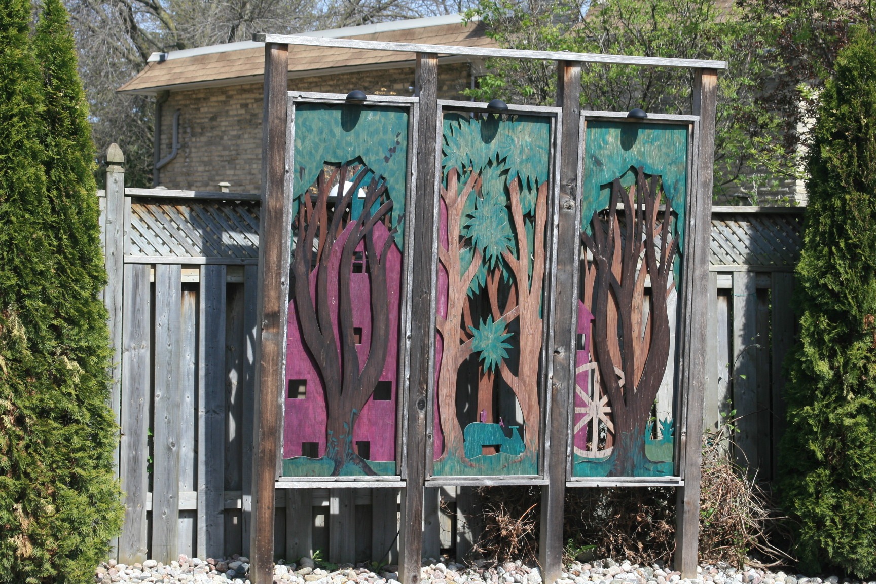 Wooden art installation with tree designs in a backyard setting, flanked by a wooden fence and evergreen shrubs under a clear sky.