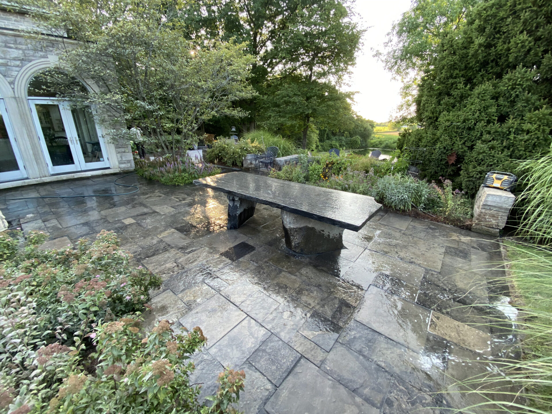 Stone patio with rectangular table, surrounded by lush greenery. Glass doors lead to a building. Peaceful garden setting under a clear sky.