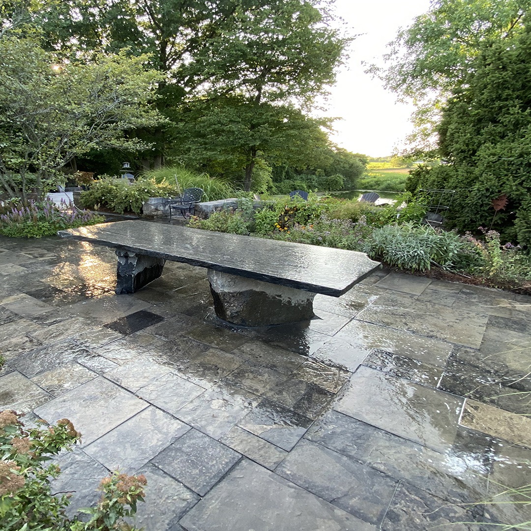 Stone patio with a sleek table, surrounded by lush greenery and trees. The serene natural setting offers a peaceful outdoor atmosphere.