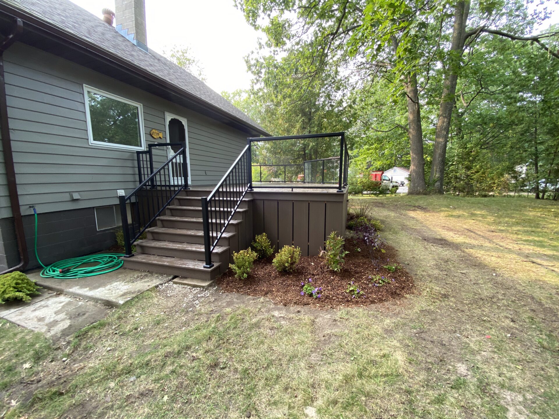 A house with gray siding features a small deck and stairs, surrounded by a grassy yard with trees and a garden bed.