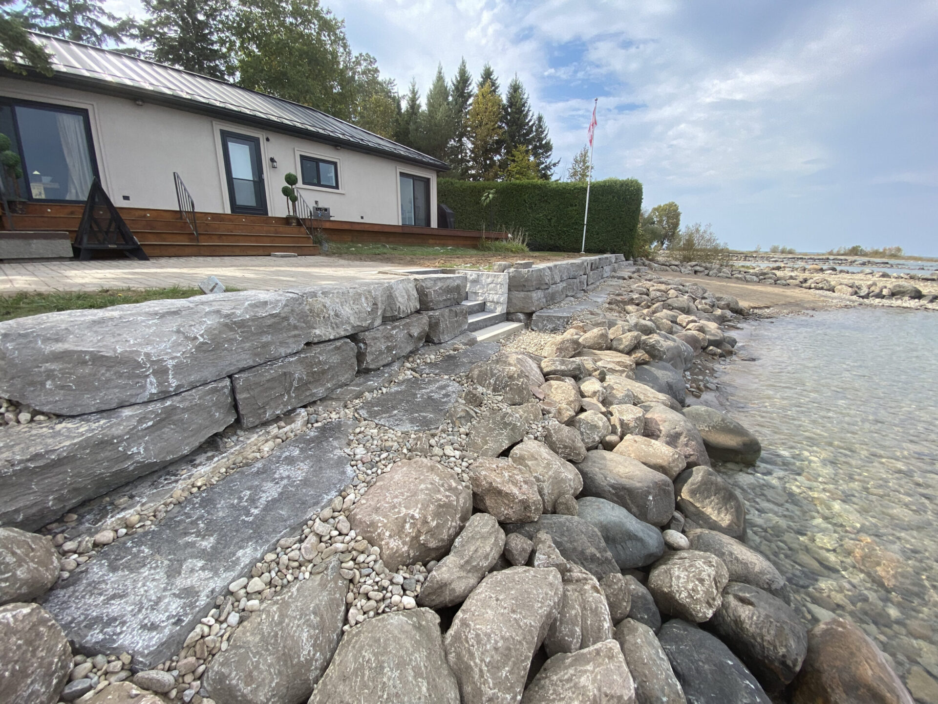 House with wooden deck overlooks rocky shoreline and water. Stone steps lead down to the water, under a cloudy sky with scattered trees.