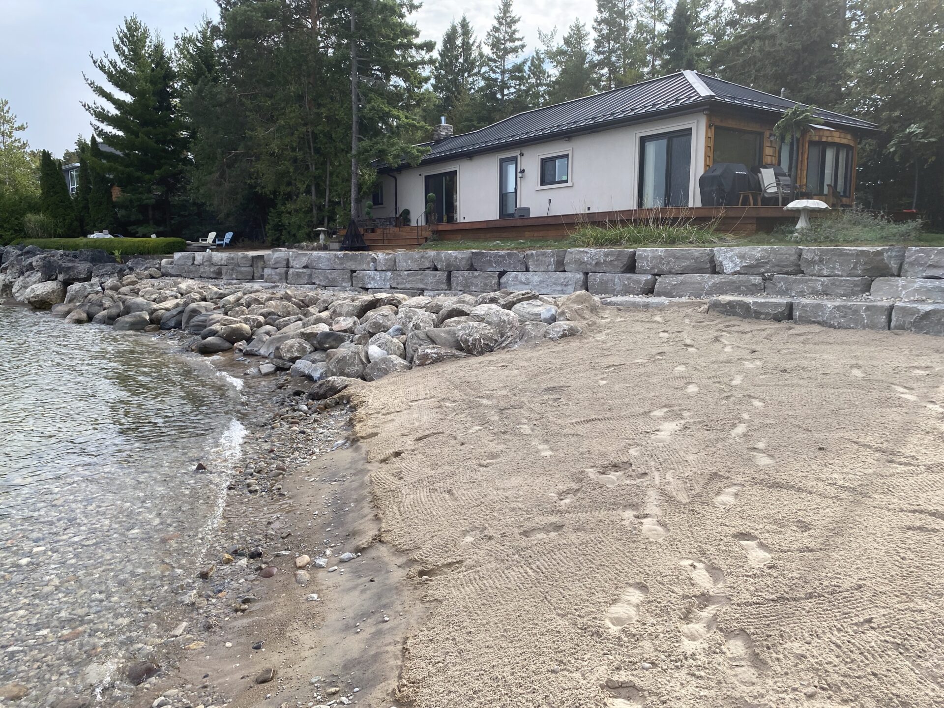 A lakeside house with a stone retaining wall, sandy beach, and surrounded by trees, under a cloudy sky. No recognizable landmarks present.