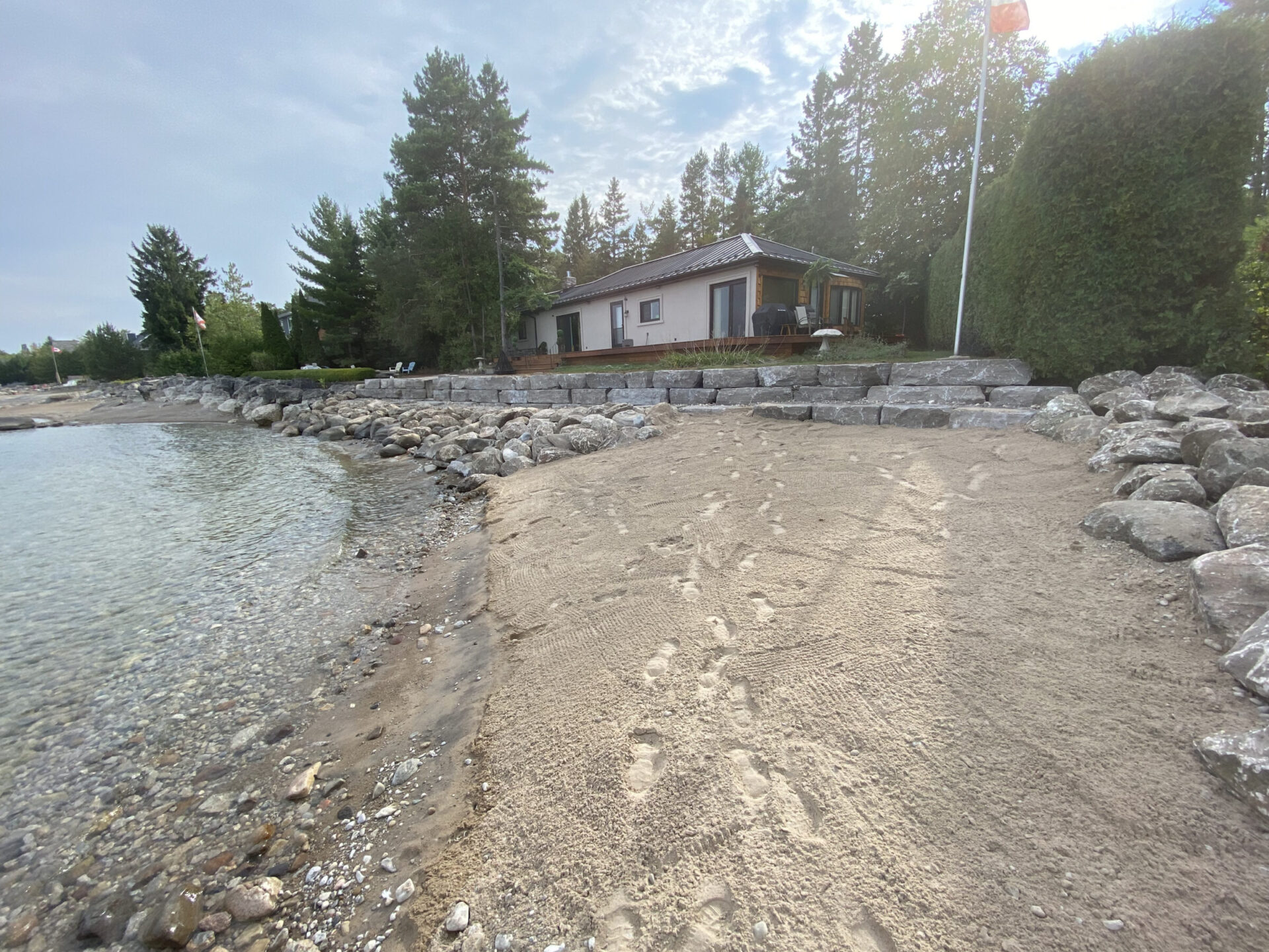 Serene lakeside scene with a modest house, sandy beach, and rock-lined shore, surrounded by lush trees under a partly cloudy sky.