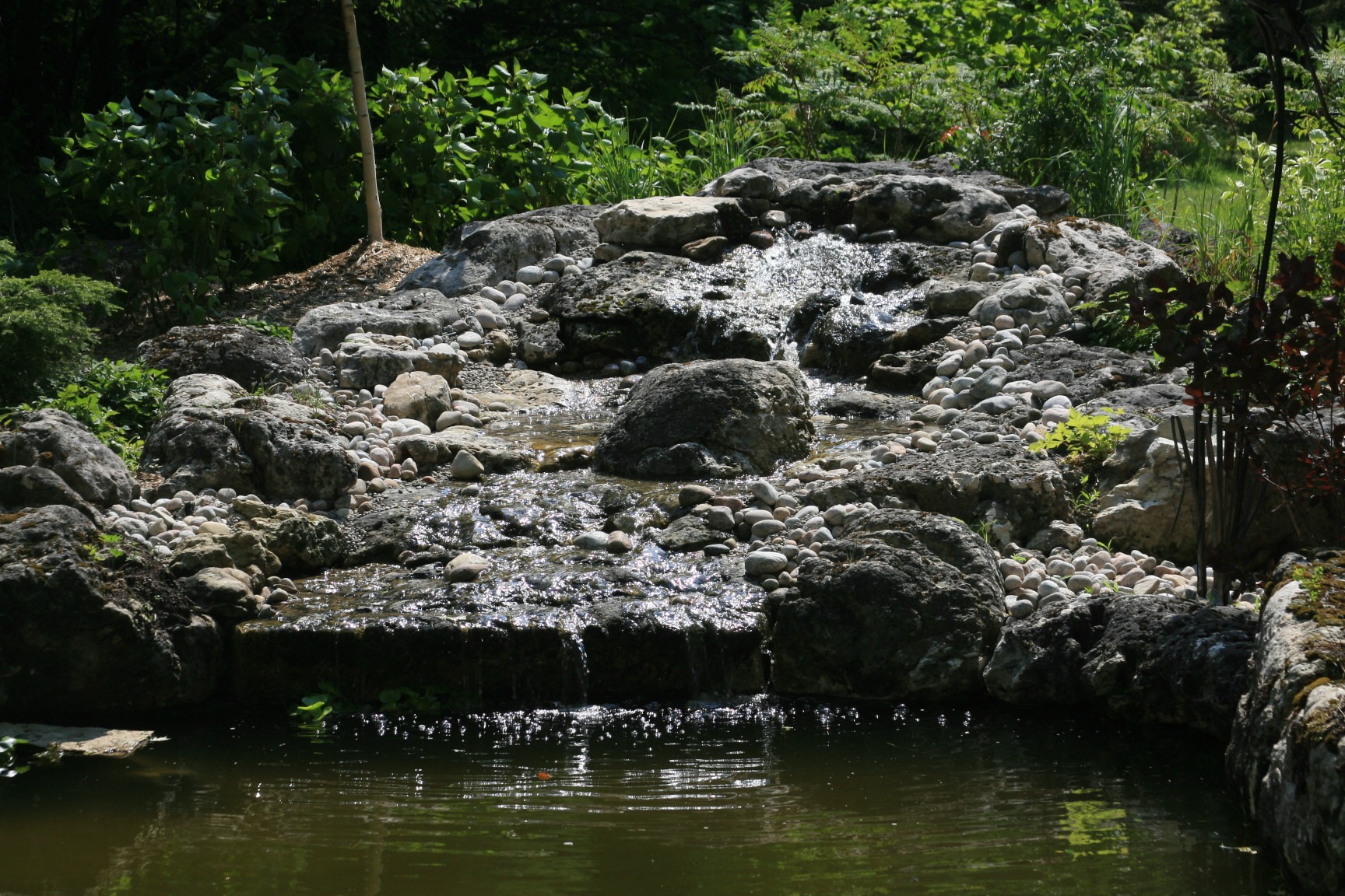 Serene garden scene with a small waterfall flowing over rocks into a tranquil pond, surrounded by lush greenery.