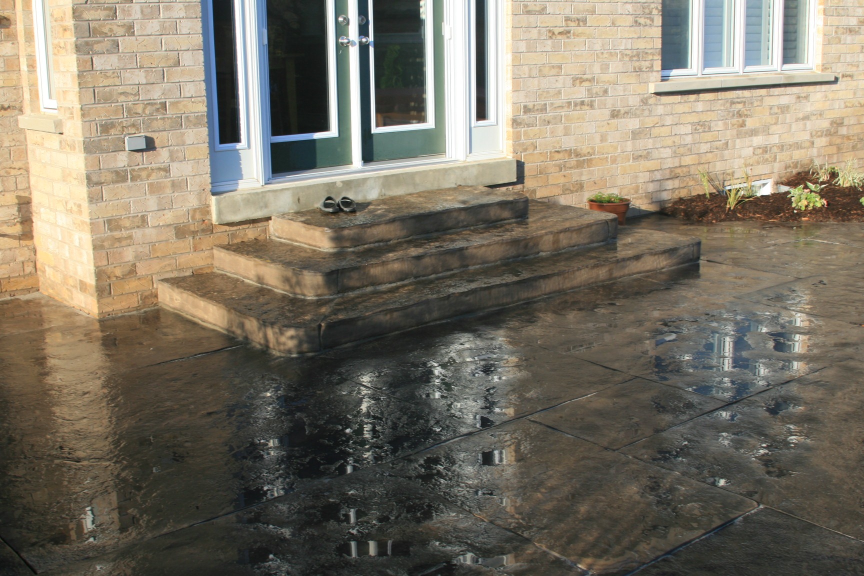 A wet concrete patio with steps leading to green double doors, bordered by a brick wall. A potted plant sits near the entrance.