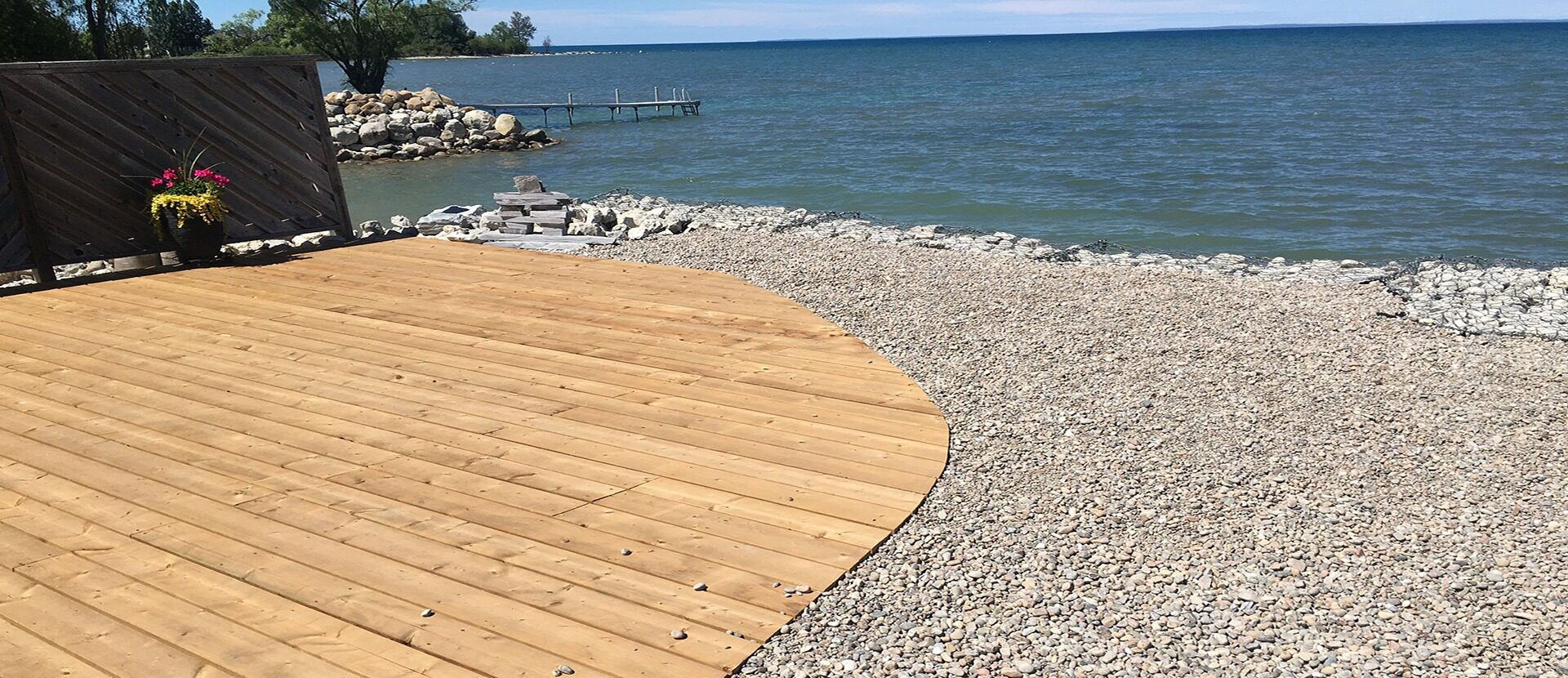 Wooden deck overlooks a rocky shoreline with a clear blue lake under a sunny sky. A flower pot decorates the scene.