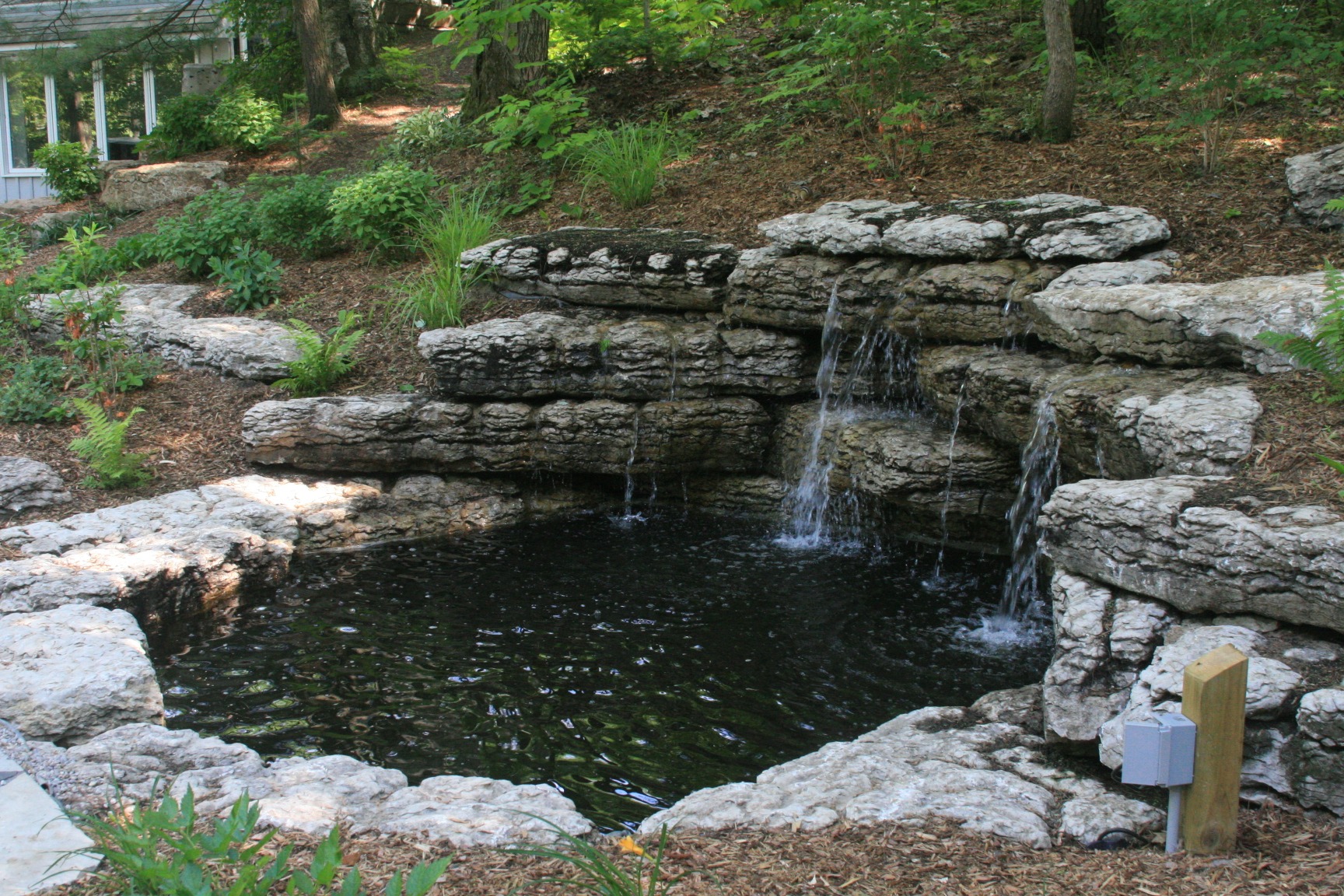 A small, naturalistic pond with cascading waterfalls surrounded by rocks and lush greenery in a tranquil, wooded garden setting.