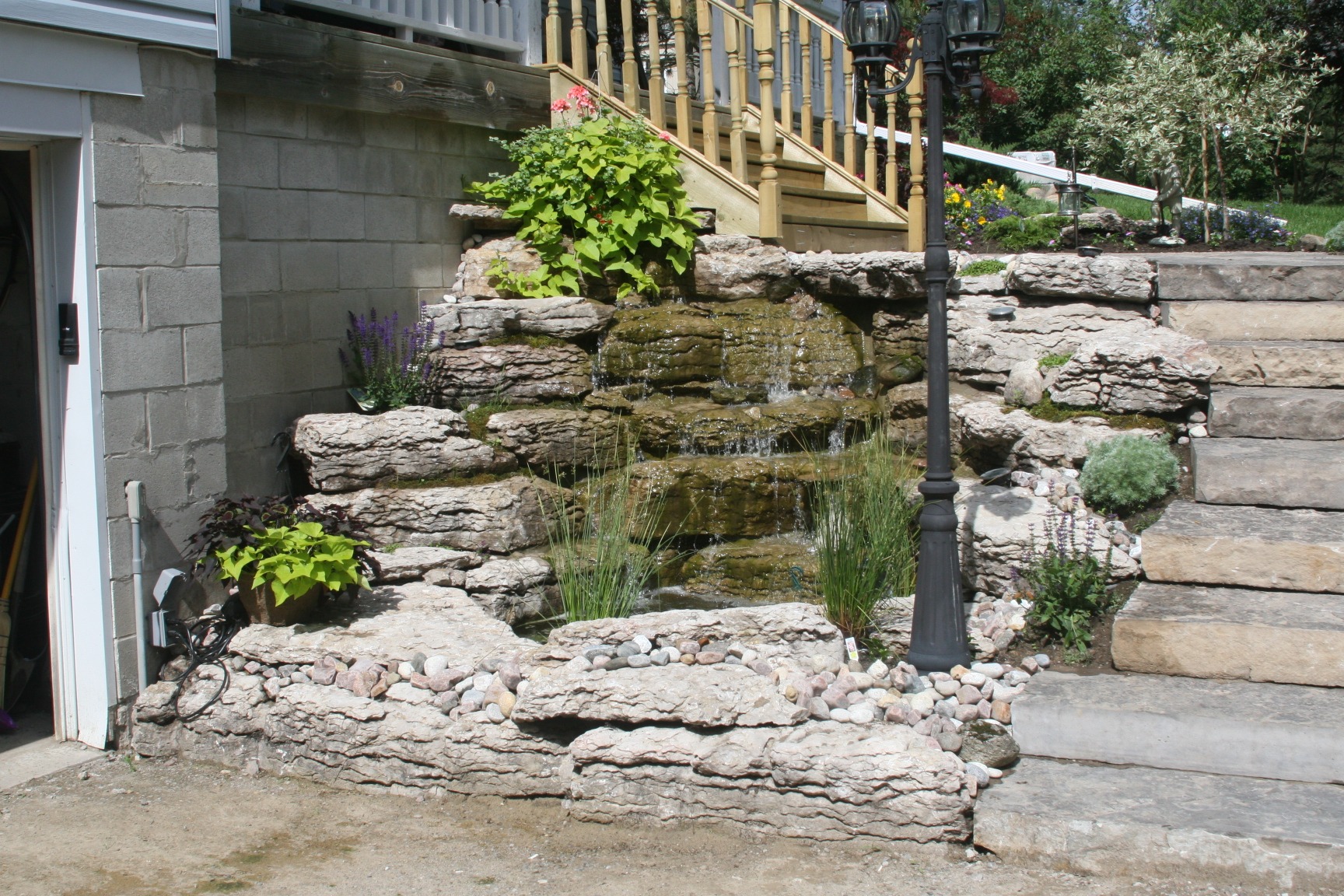 A small garden waterfall with stone steps, greenery, and a black lamppost. Natural and serene backyard landscape design near a building.