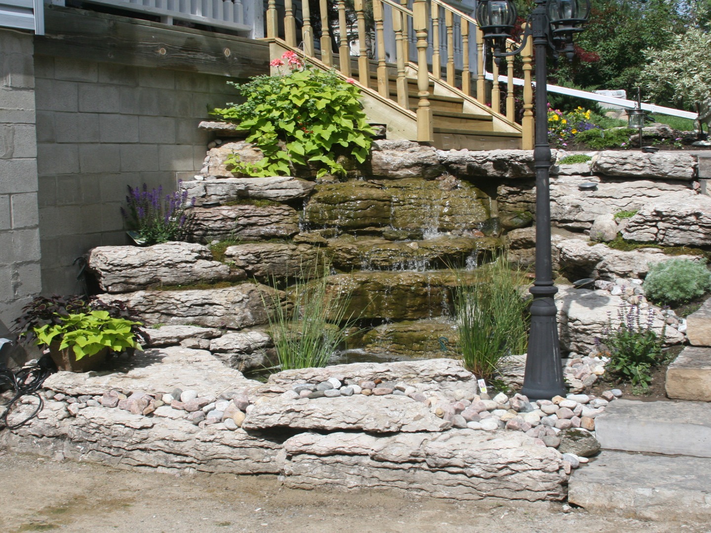 Stone garden waterfall with layered rocks, surrounded by plants and pebbles. A lamppost stands nearby, next to wooden stairs and greenery.