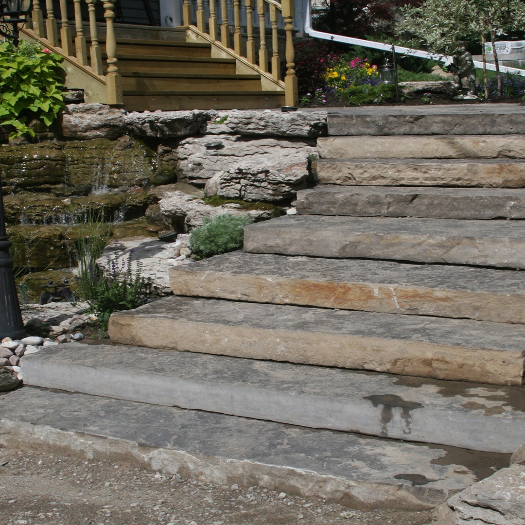 A stone staircase leads to a porch with yellow railings. Natural rocks and plants decorate the sides, creating a rustic outdoor setting.