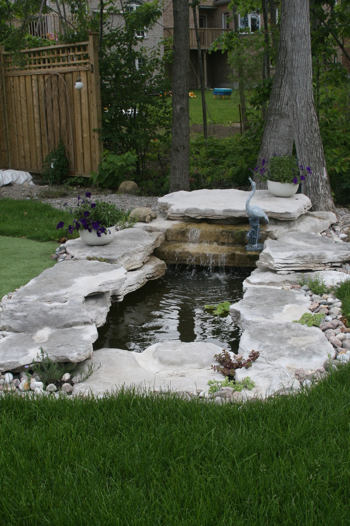 A tranquil backyard garden features a small stone fountain with cascading water, surrounded by lush greenery and decorative plants.