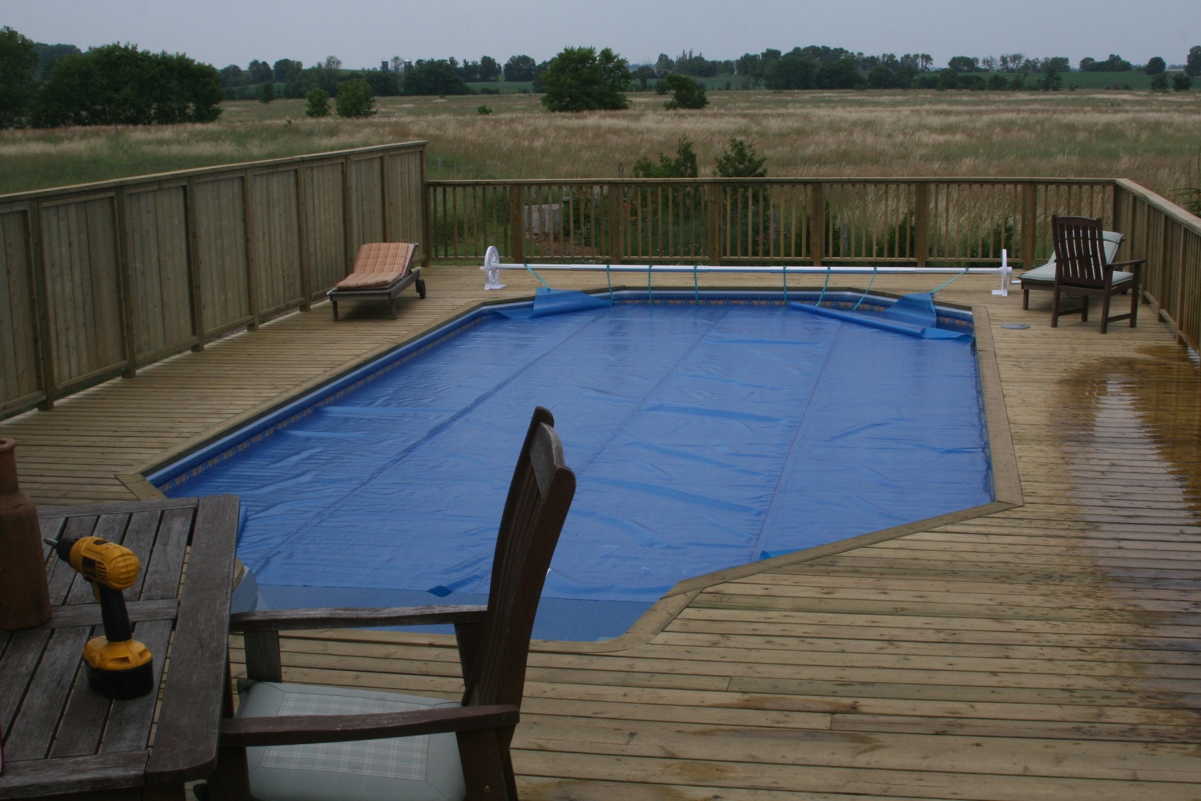 A wooden deck surrounds a covered outdoor pool with chairs and a table, set against a backdrop of open fields and trees.