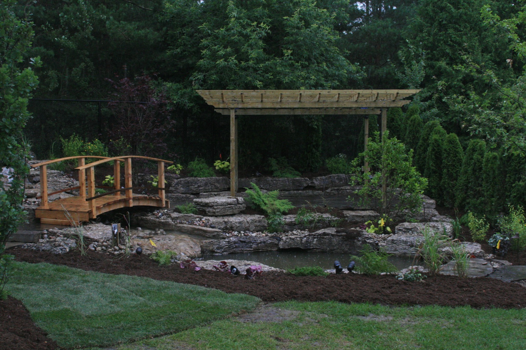 A serene garden features a wooden bridge, pergola, stone-lined pond, and lush greenery, creating a tranquil, natural setting surrounded by trees.