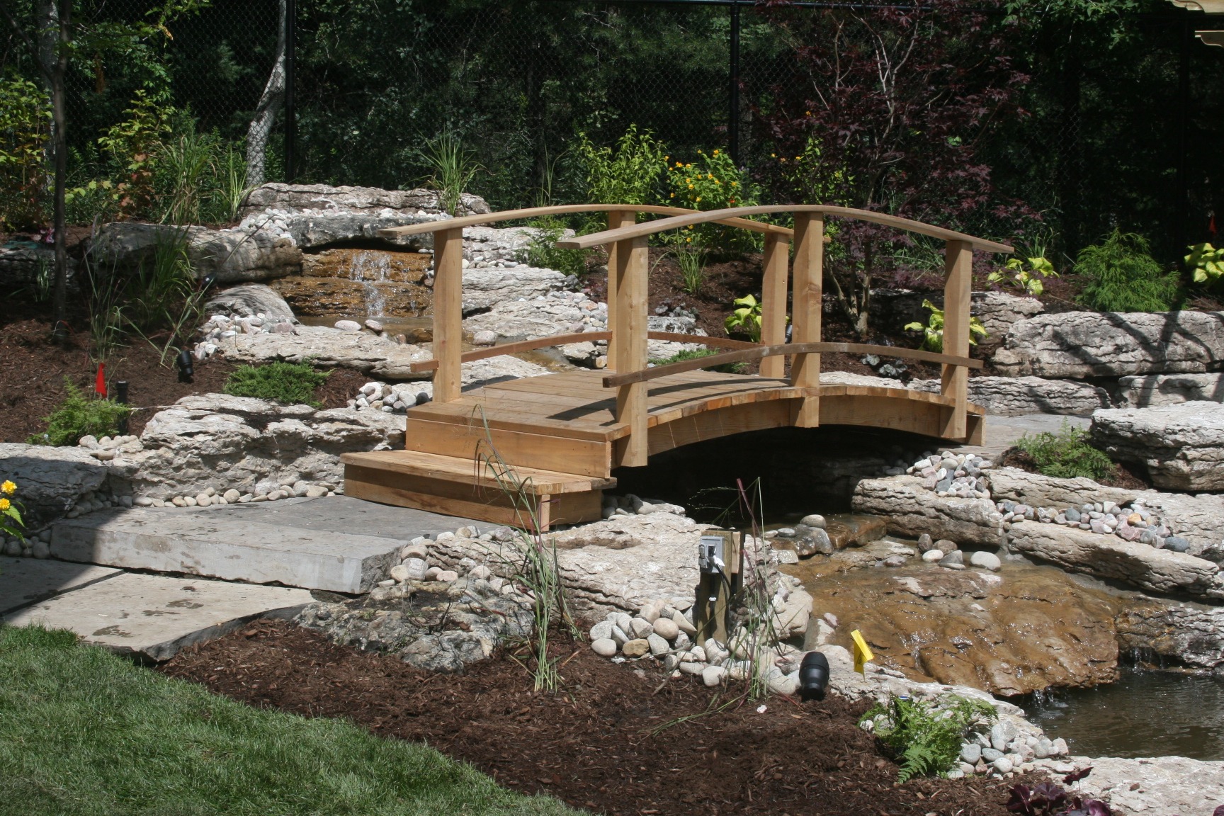 A wooden footbridge spans a small garden pond surrounded by rocks and greenery, creating a tranquil natural landscape scene.