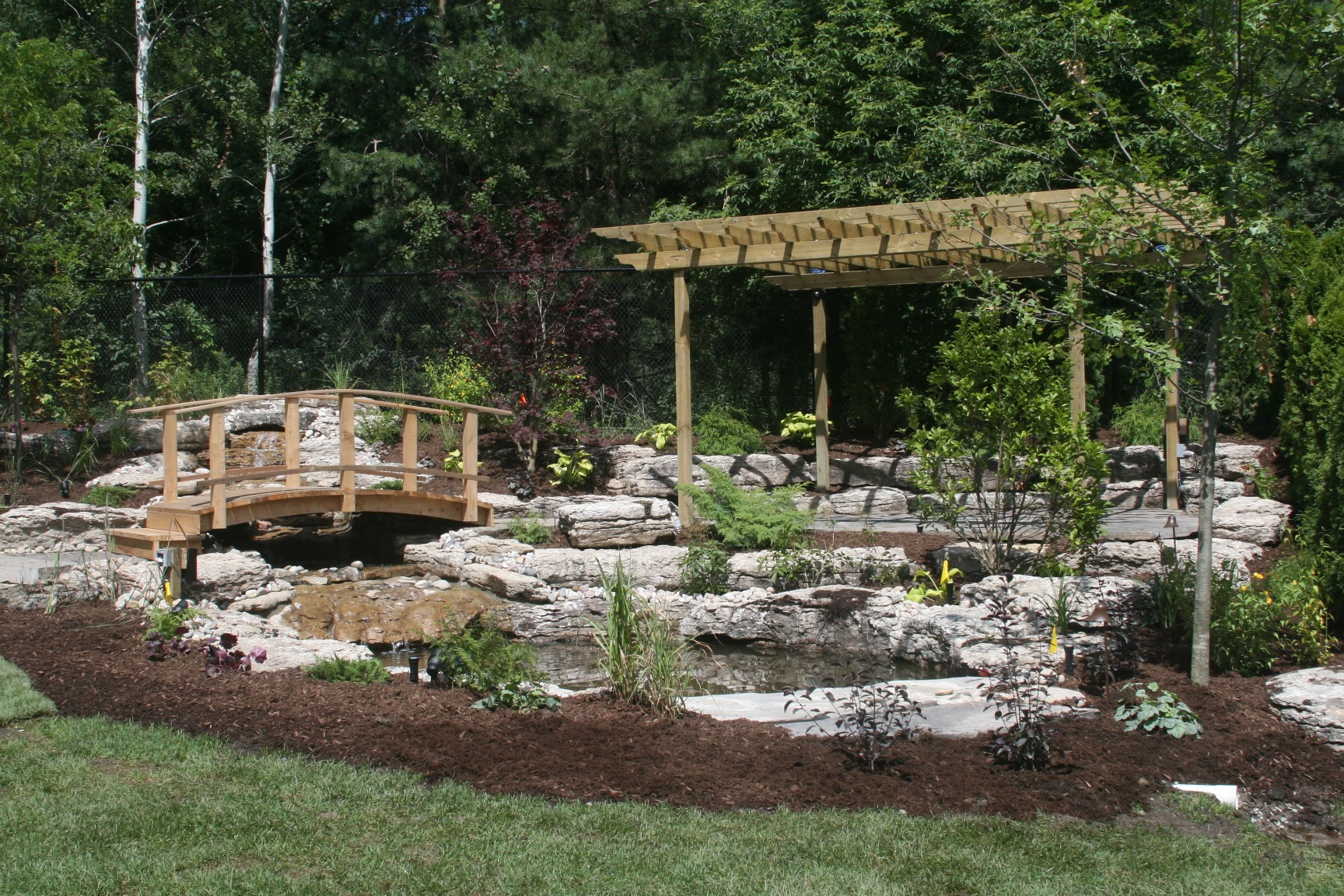 A serene garden with a wooden bridge over a water feature, lush greenery, and a pergola, surrounded by rocks and trees.