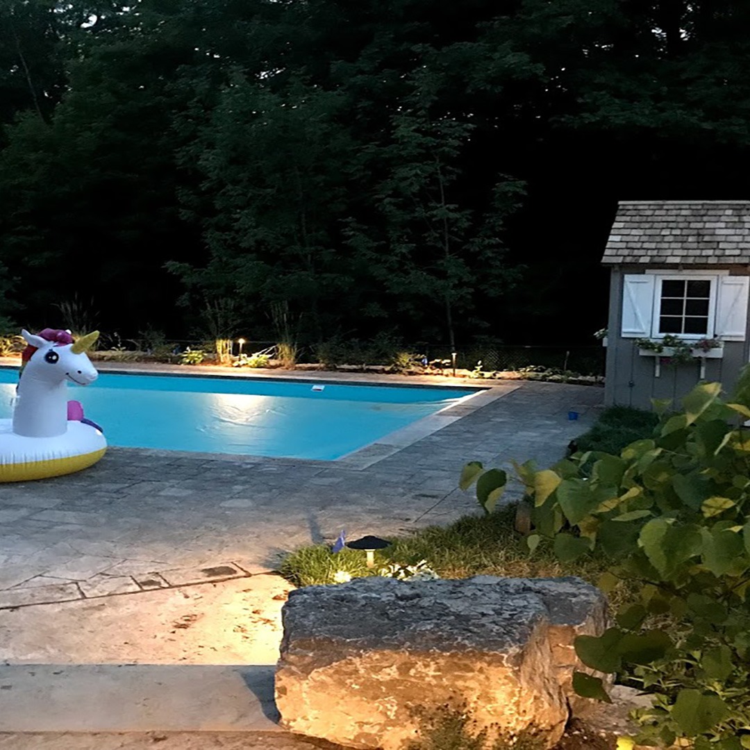 A backyard pool with a unicorn float, surrounded by trees and small shed, creates a serene evening atmosphere with soft lighting.