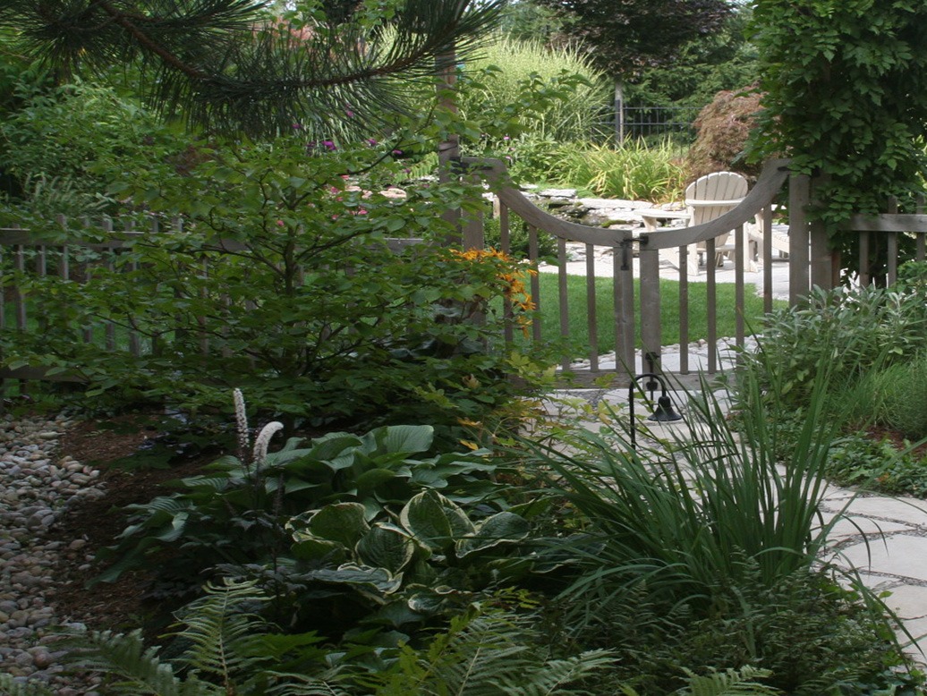 A lush garden with diverse plants, a curved wooden gate, and a view of a patio with white chairs in the background.