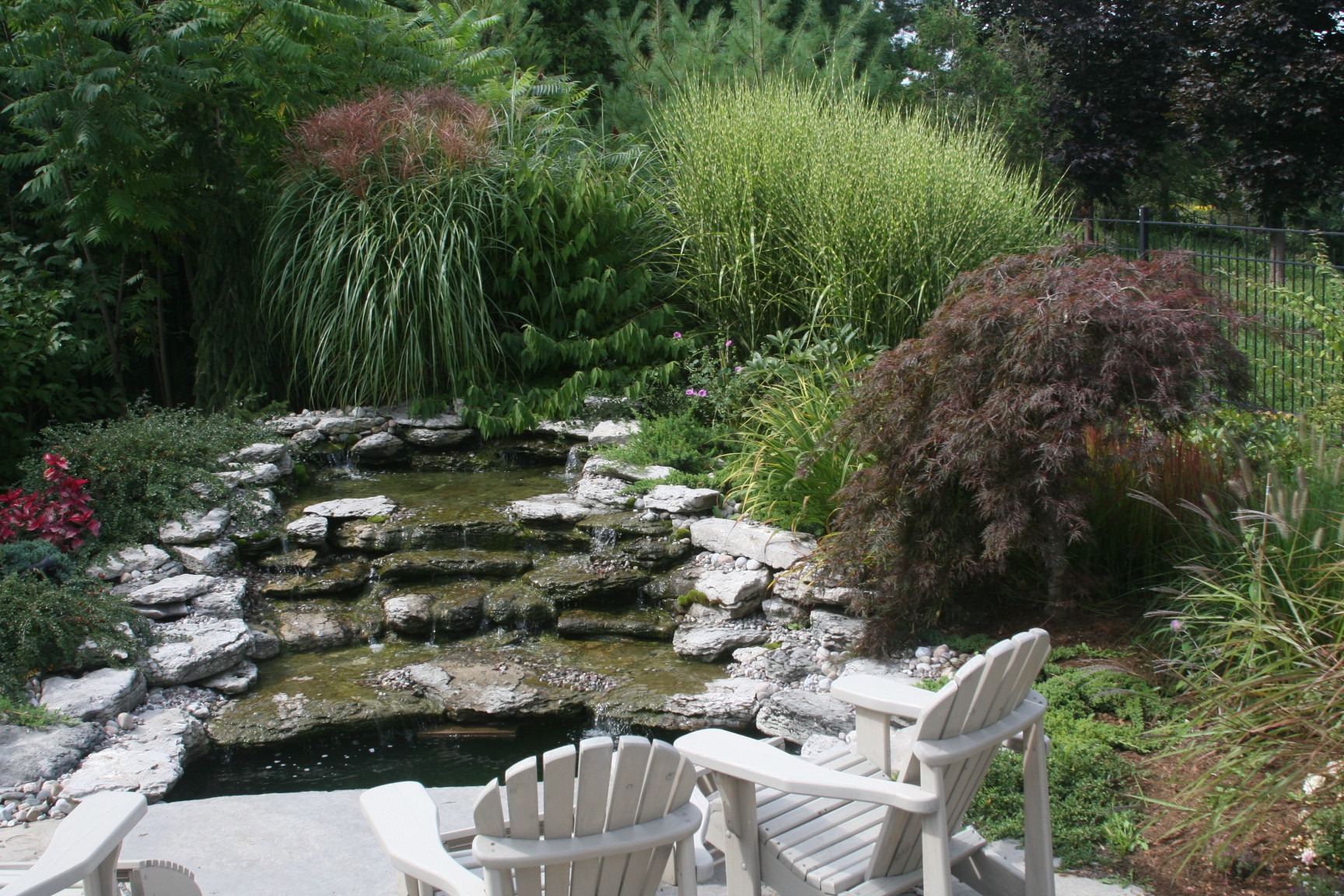 A serene garden scene with a small cascading waterfall, surrounded by lush greenery and Adirondack chairs on a stone patio.