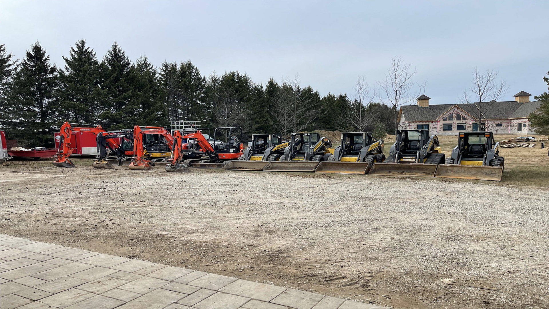 A lineup of construction vehicles, including excavators and loaders, on a gravel lot with trees and a large building in the background.