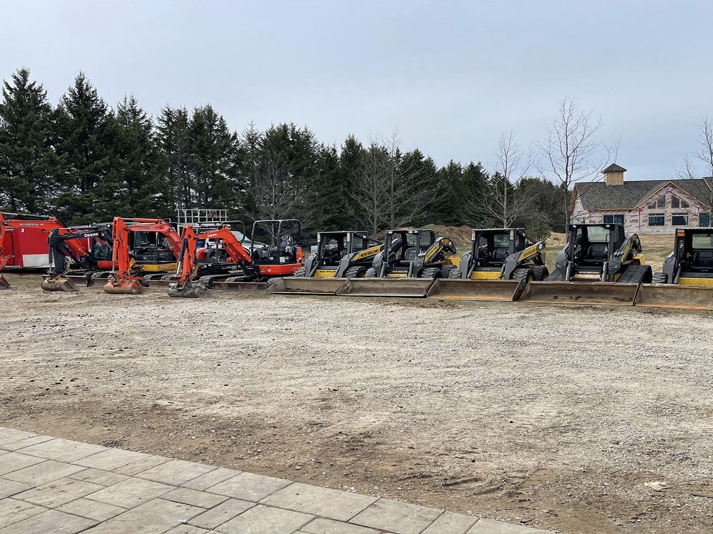 A row of construction vehicles, including excavators and loaders, parked on a gravel lot with trees and a building in the background.