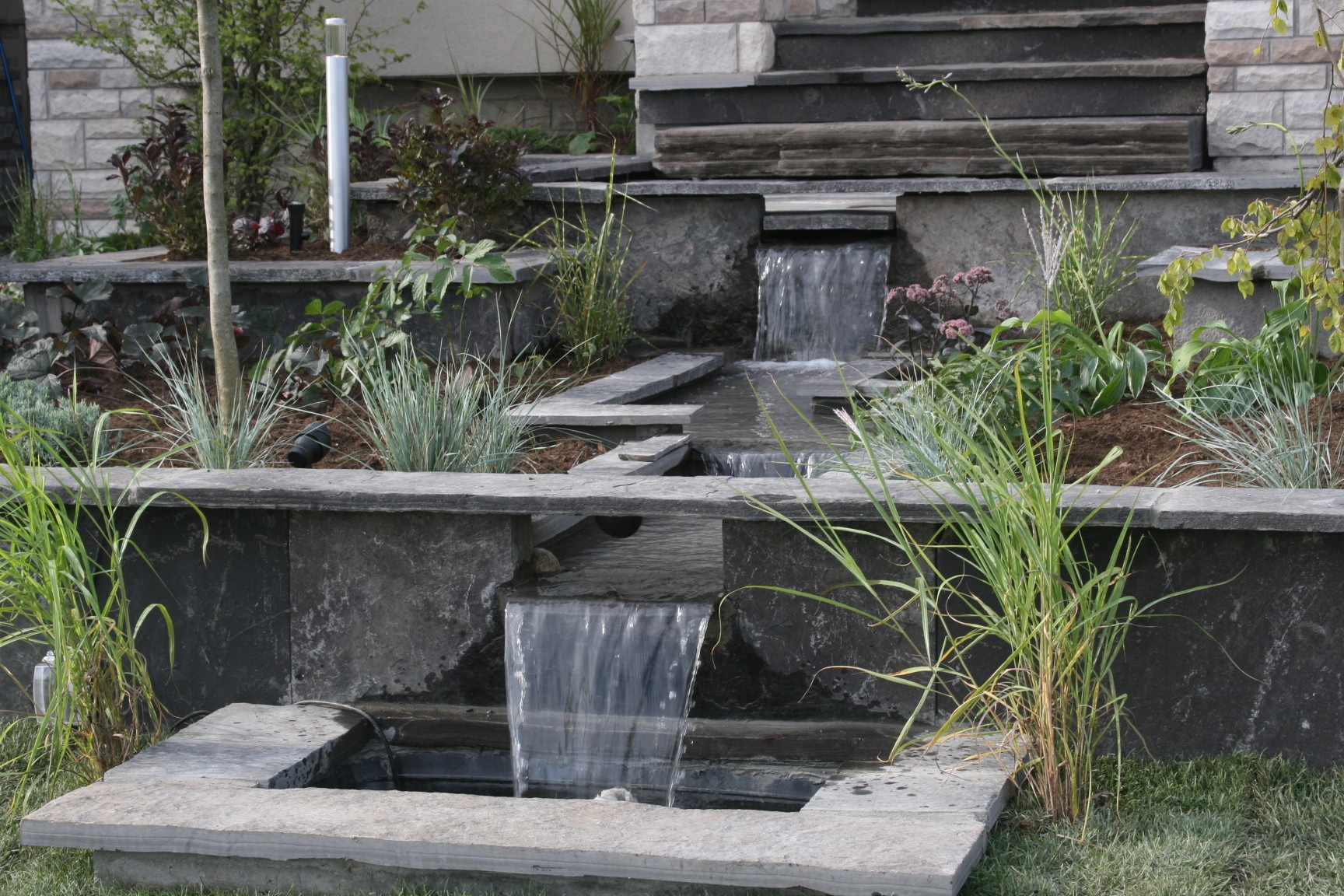 A tiered stone water feature in a garden, surrounded by plants and shrubs, cascading water creating a tranquil ambiance.