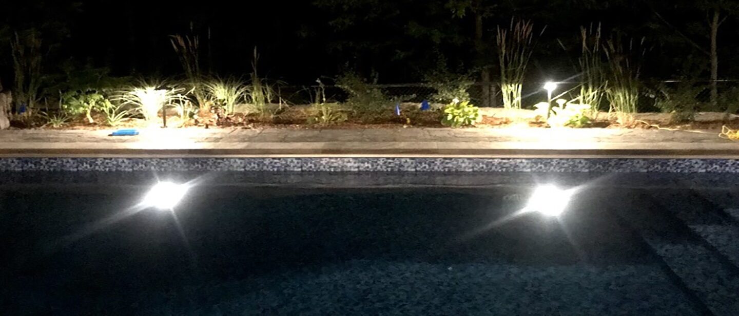 A dimly lit outdoor pool at night, with glowing lights illuminating a landscaped garden area against a backdrop of trees.