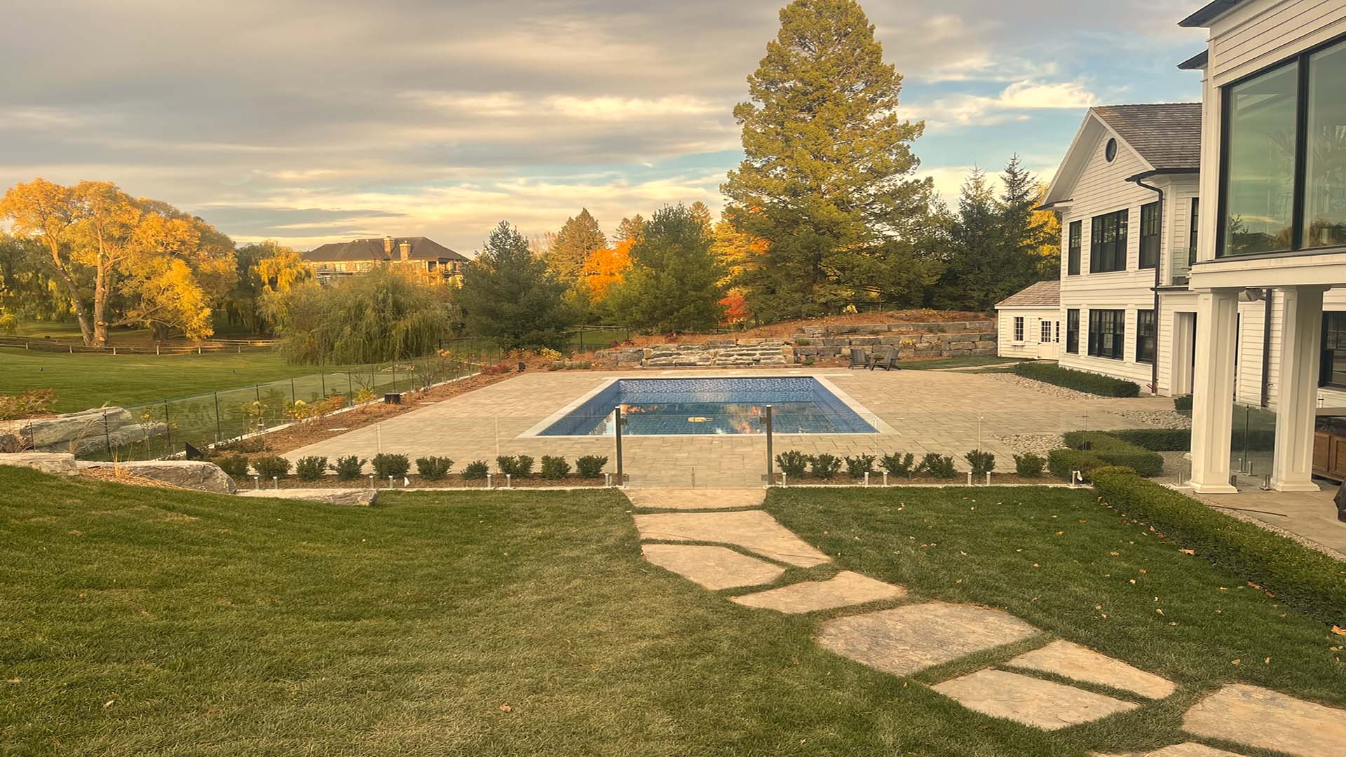 A spacious backyard features a rectangular swimming pool, surrounded by a well-manicured lawn and trees, leading to a large, modern house.