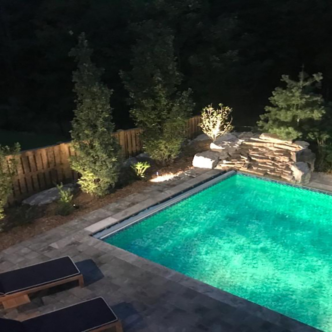 A nighttime view of a well-lit swimming pool with surrounding trees and lounge chairs, enclosed by a wooden fence and landscaped rocks.