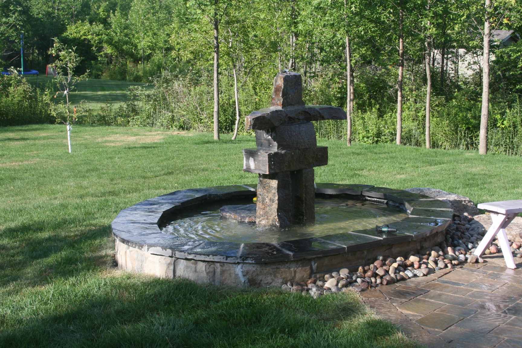 A backyard scene featuring an inukshuk-style stone fountain on a circular platform. Surrounding greenery includes trees and grass under a clear sky.