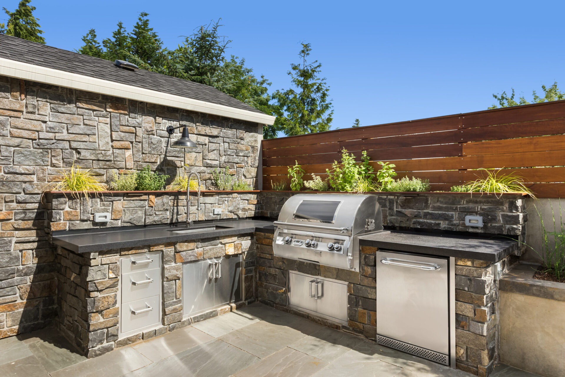 Outdoor kitchen setup with stainless steel grill, appliances, and stone countertops. Wooden privacy fence and greenery in the background. Bright, clear day.