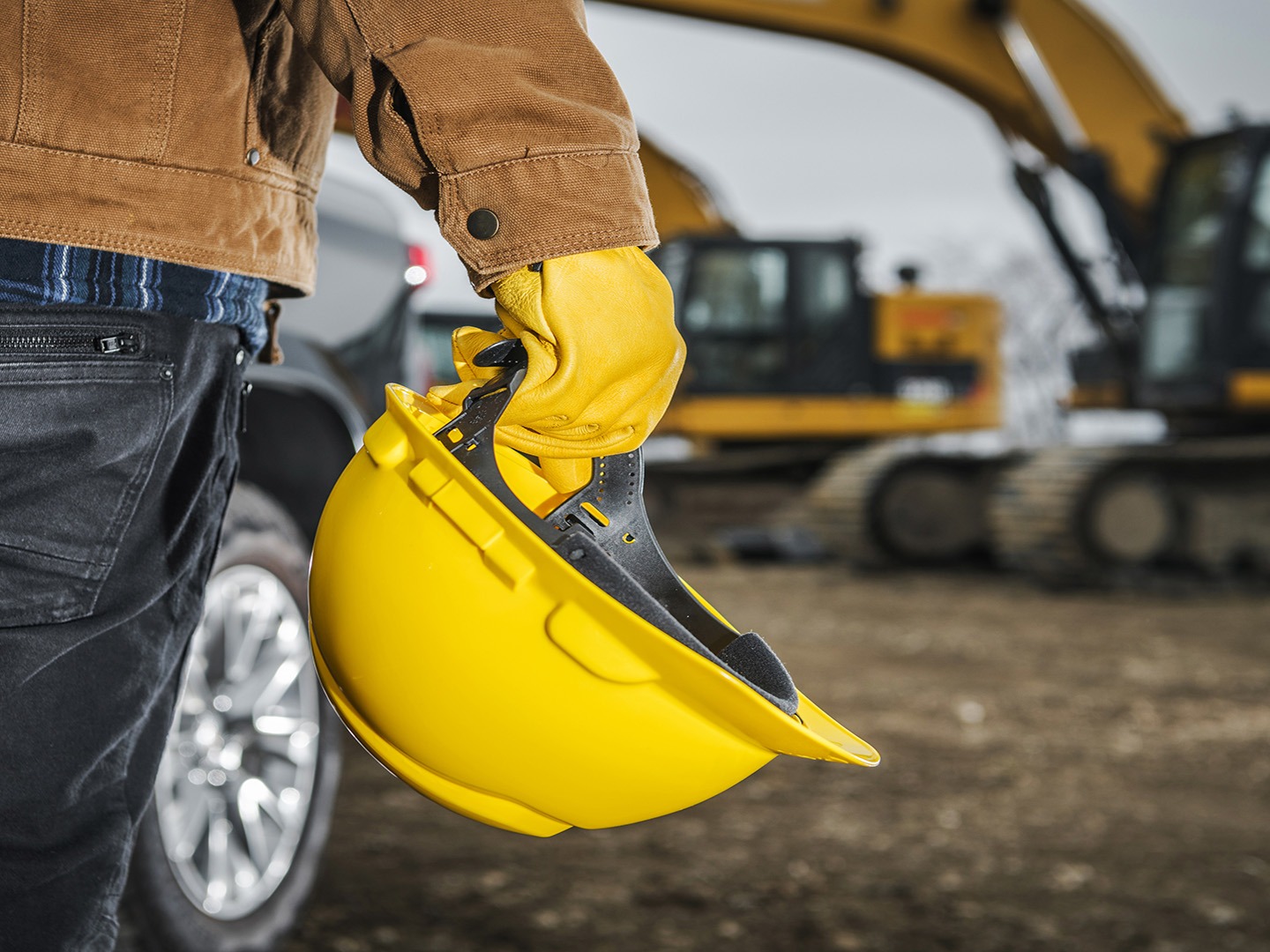 A person holding a yellow hard hat stands near construction equipment on a dirt site, wearing a brown jacket and yellow gloves.