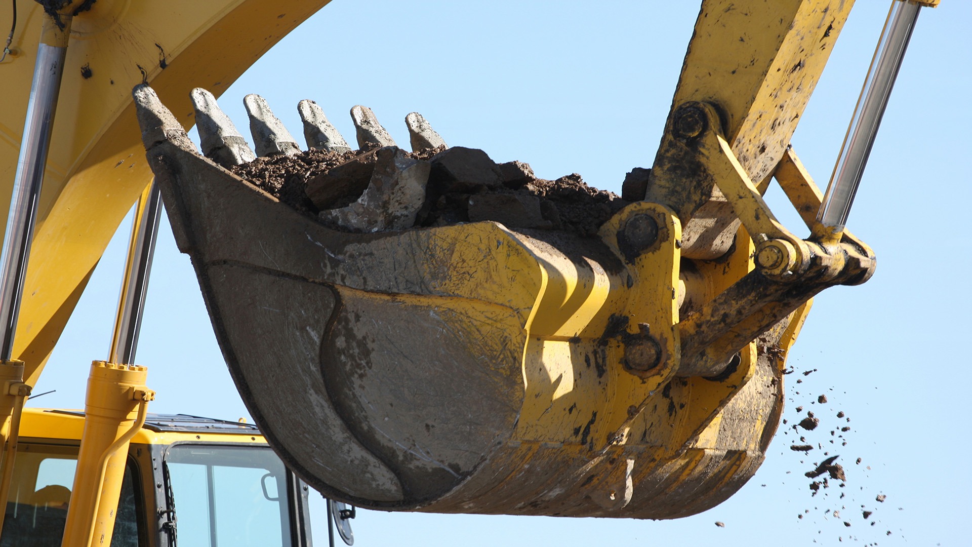 A yellow excavator scoops up dirt against a clear blue sky. The machinery's bucket is visible, releasing soil and debris.