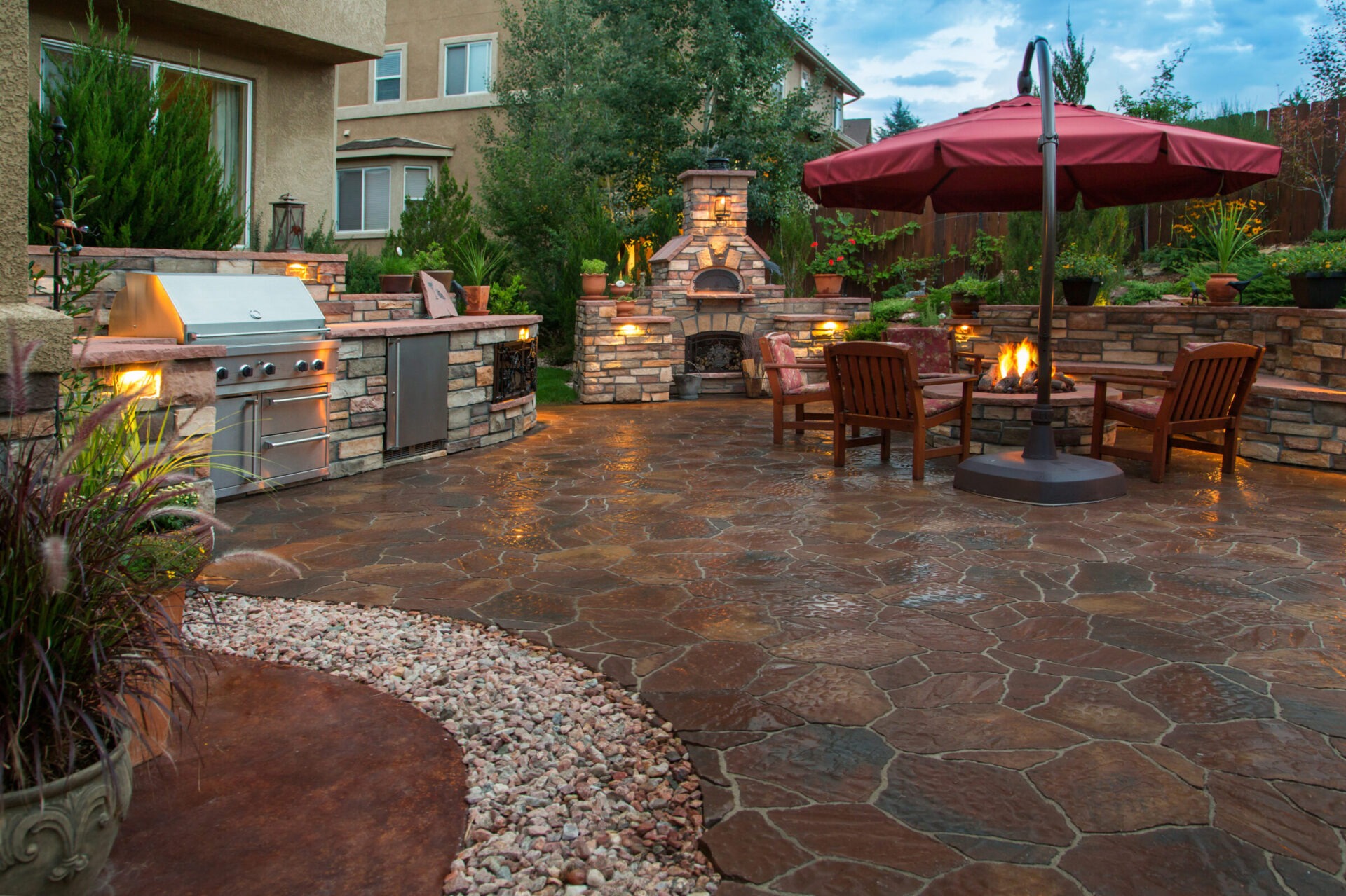 A charming backyard features a stone patio with a grill, fireplace, seating area, and umbrella, surrounded by lush greenery and soft lighting.