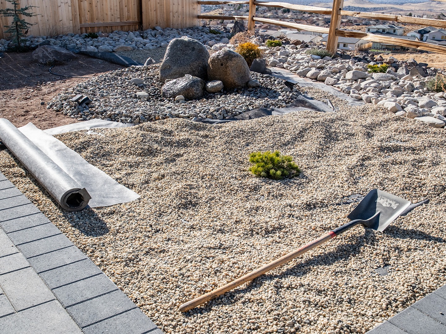 A landscaped backyard with gravel, stones, and a shovel, surrounded by a wooden fence. A rolled-up mat lies on the ground.