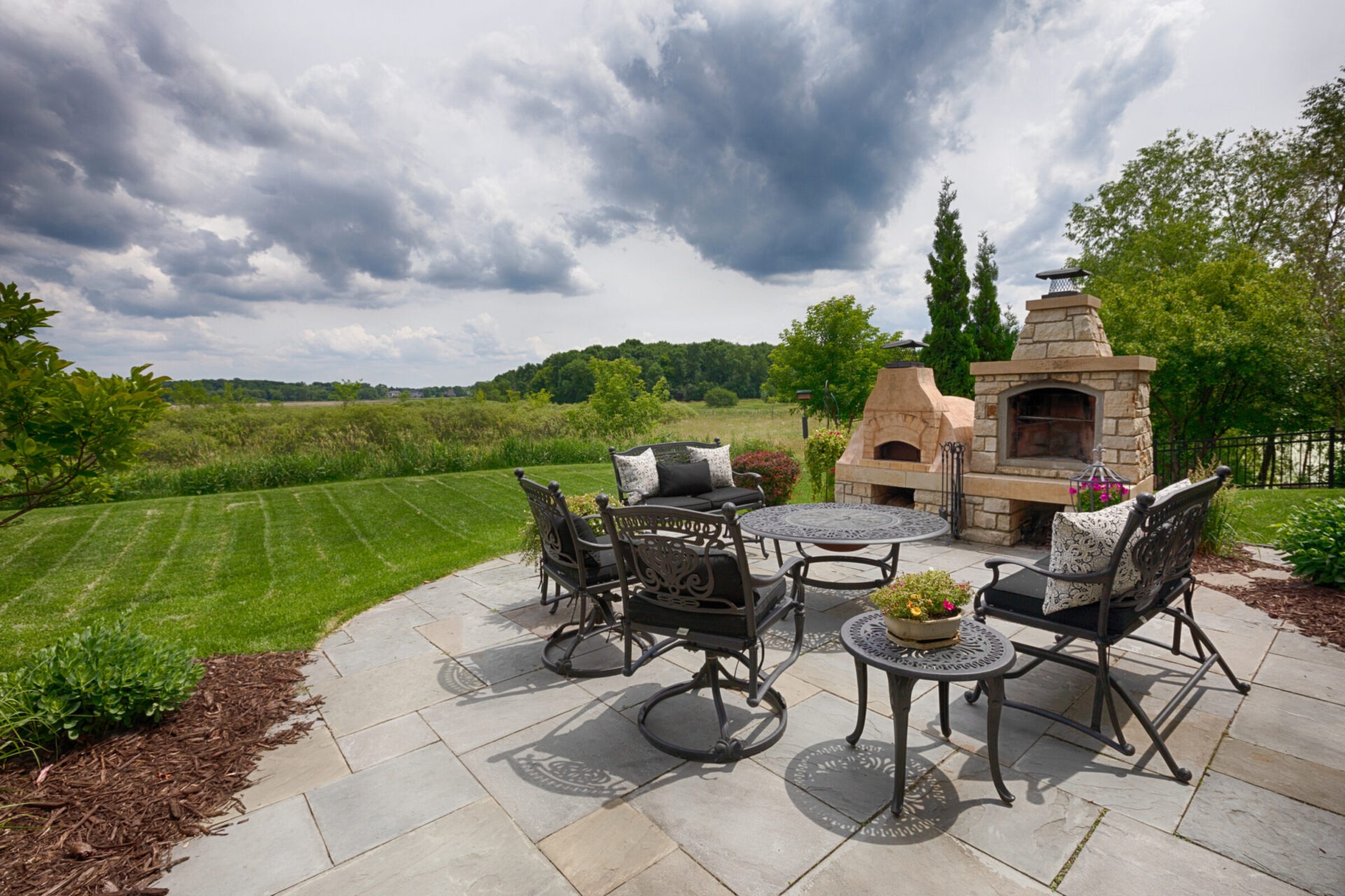 A scenic patio with metal furniture, a stone fireplace, and lush greenery under a cloudy sky. Great outdoor area for relaxation and gatherings.