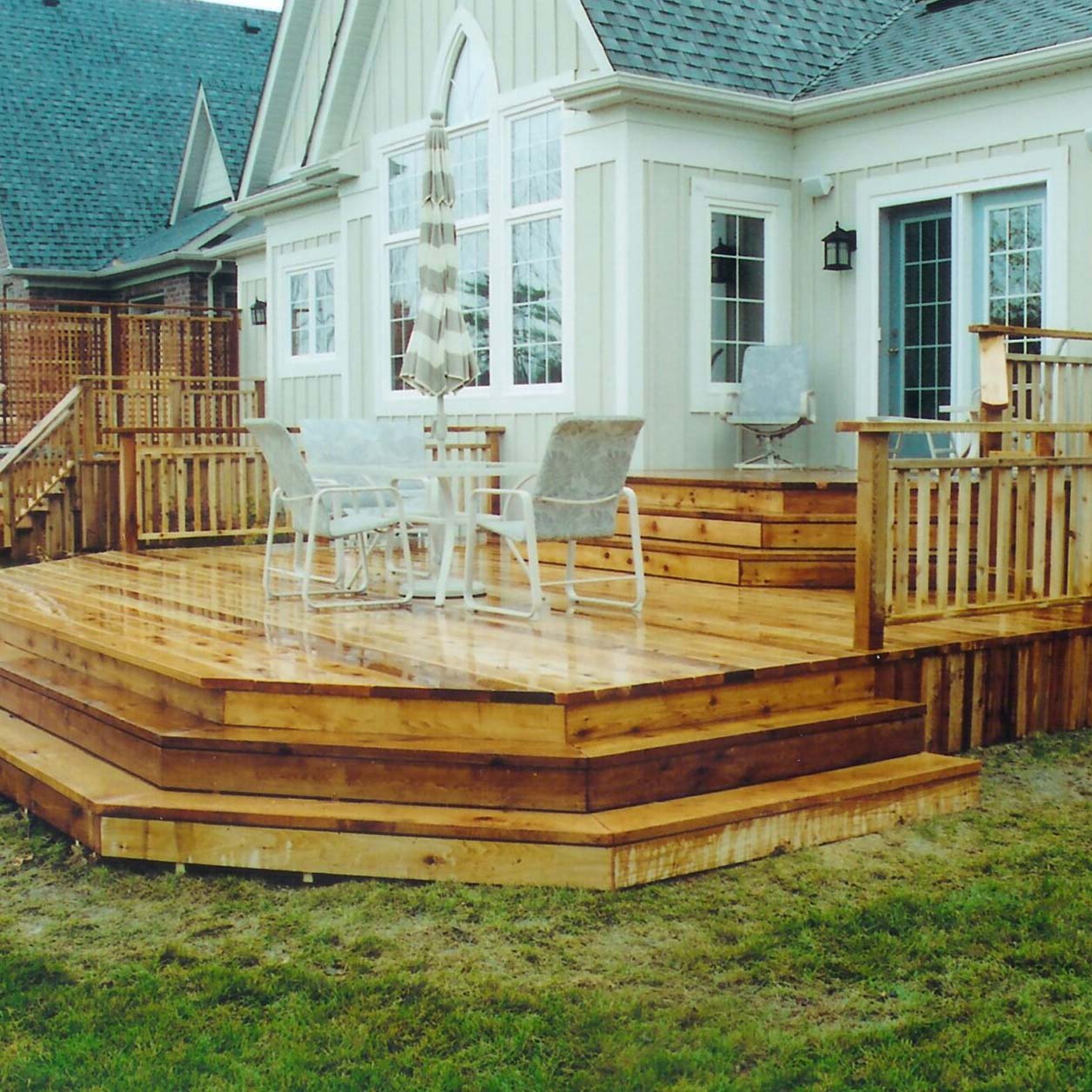 A wooden deck with table and chairs is attached to a house, featuring white siding and multiple steps leading down to the grass.