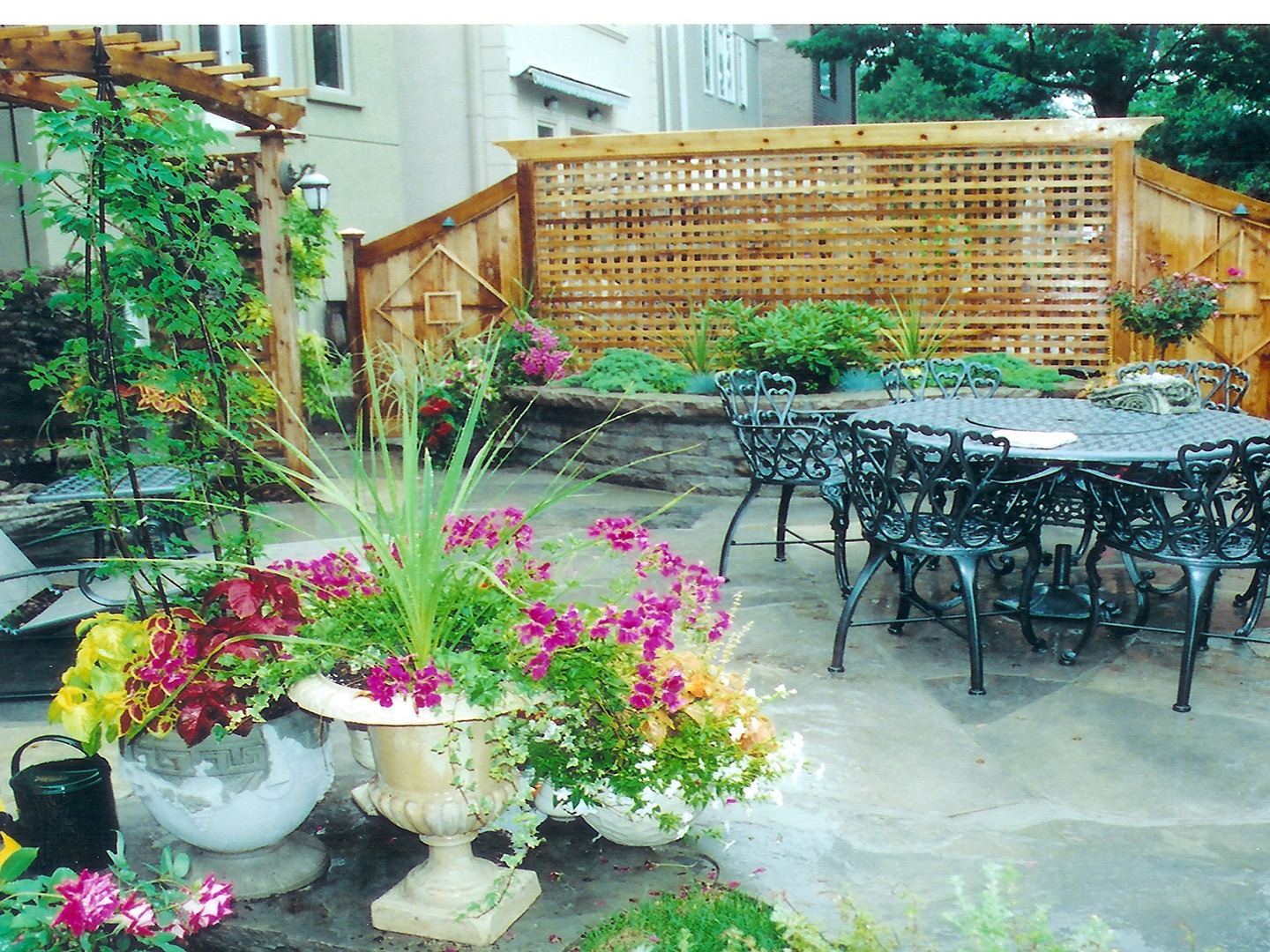 A charming garden patio features wrought iron furniture, colorful flowers in planters, and a wooden trellis creating an inviting outdoor space.