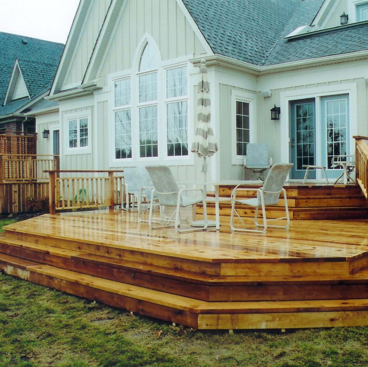 A wooden deck with chairs and an umbrella is attached to a house with large windows and tri-paneled doors, surrounded by a grassy yard.