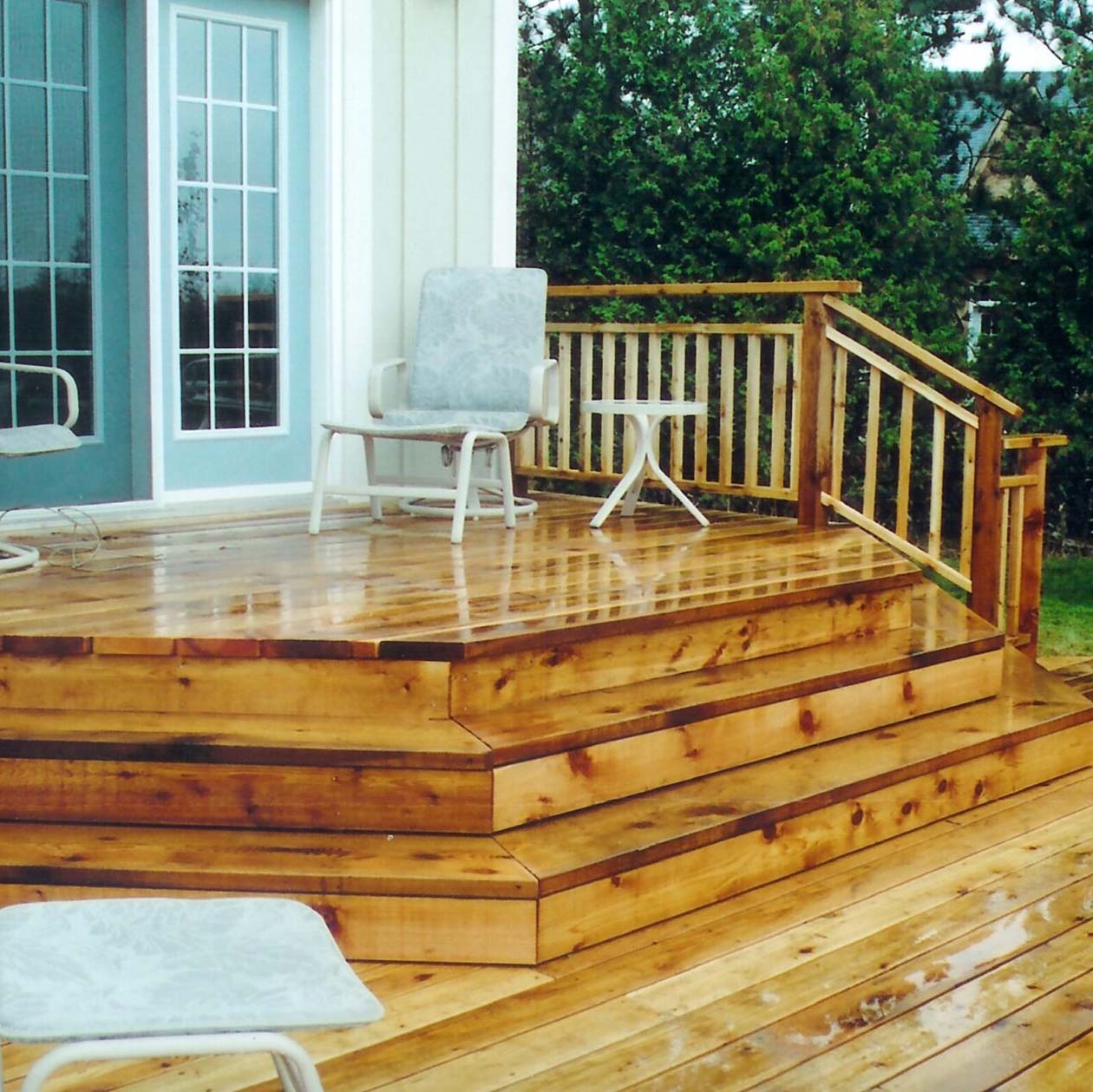 A wooden deck with steps leads to a house entrance. Outdoor chairs and small table. Surrounding greenery provides a serene atmosphere.