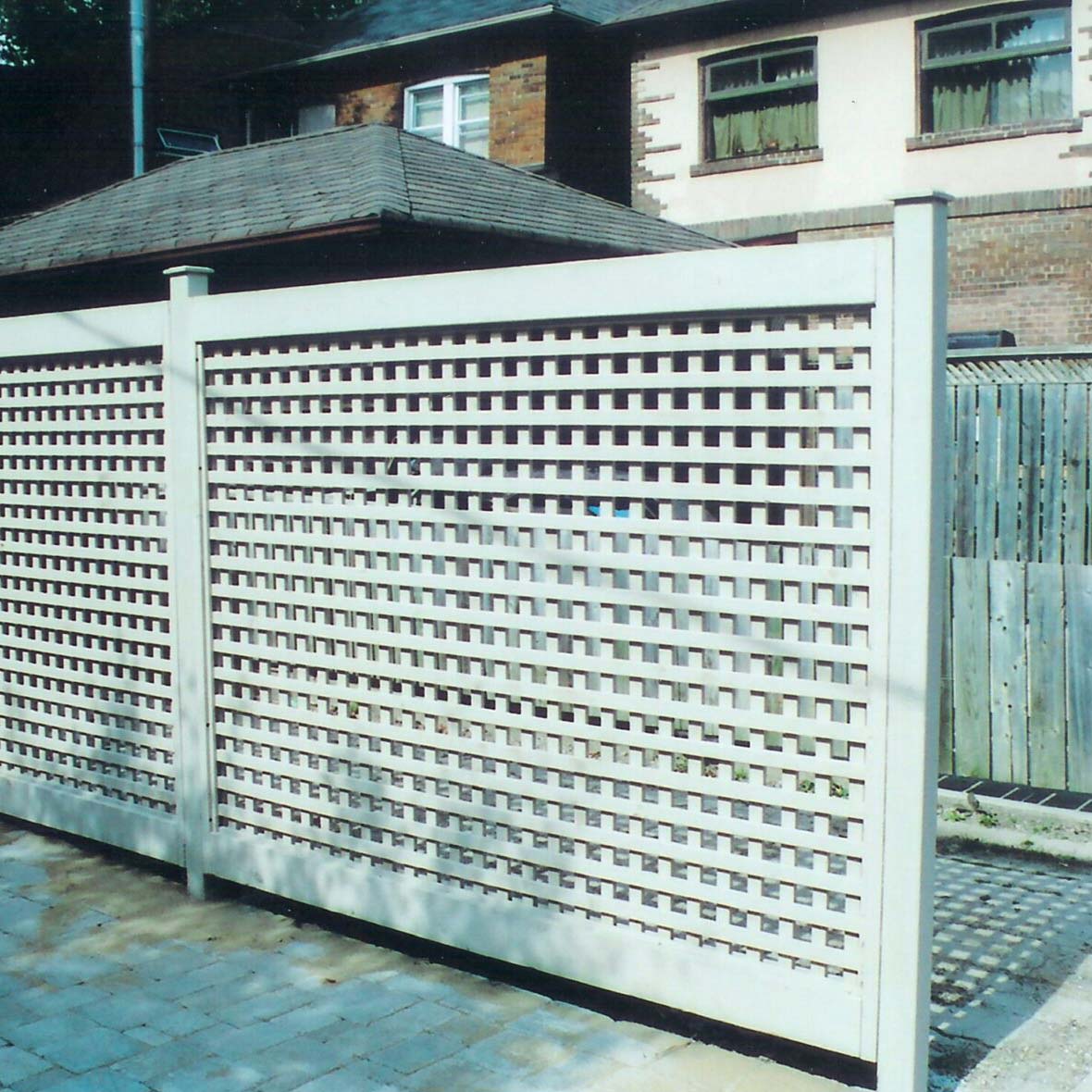 A white lattice fence stands in front of residential buildings, casting shadows on a stone-paved ground. No landmarks or historical buildings visible.