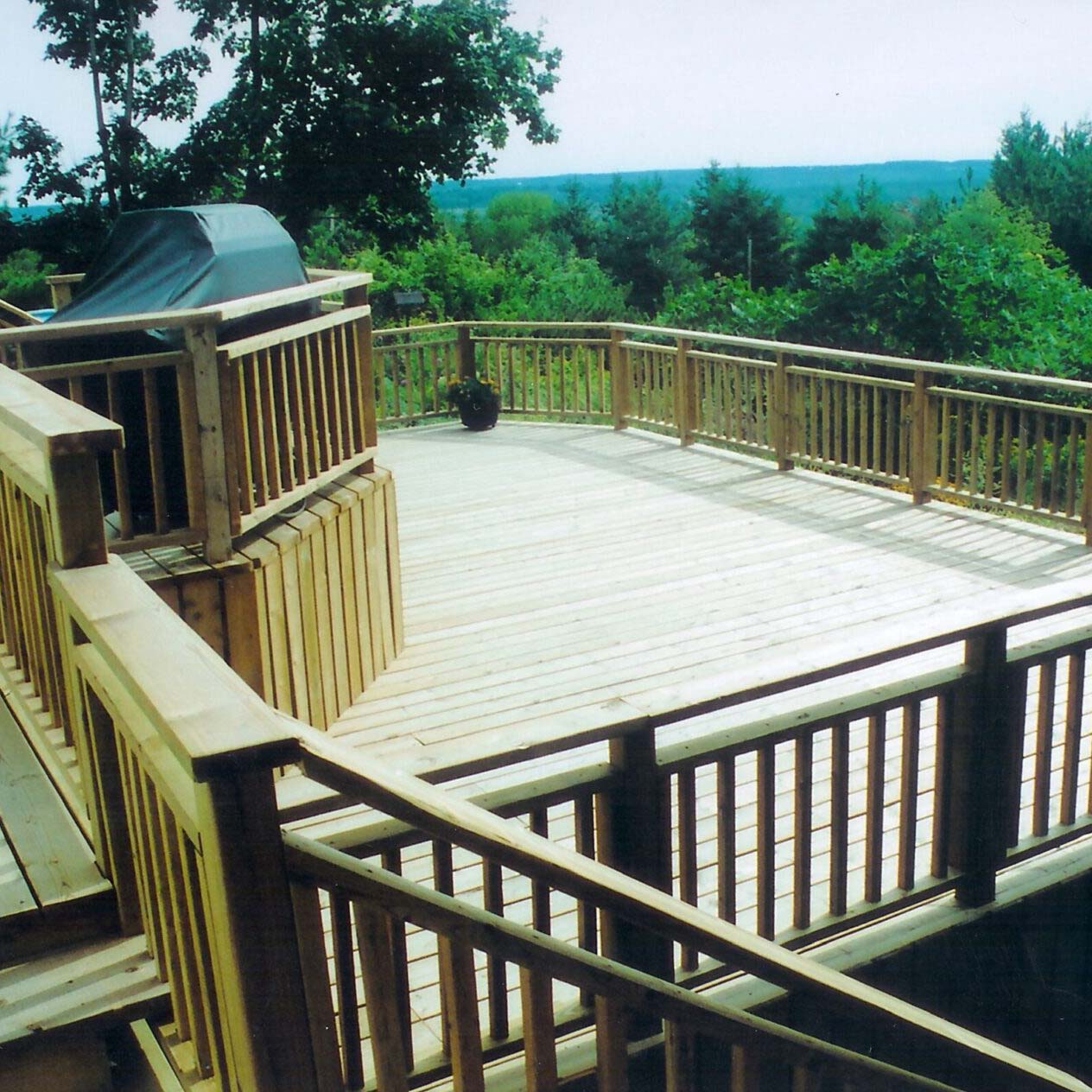 A spacious wooden deck overlooks a lush green forest. A barbecue grill is placed on the deck, surrounded by railing and trees.