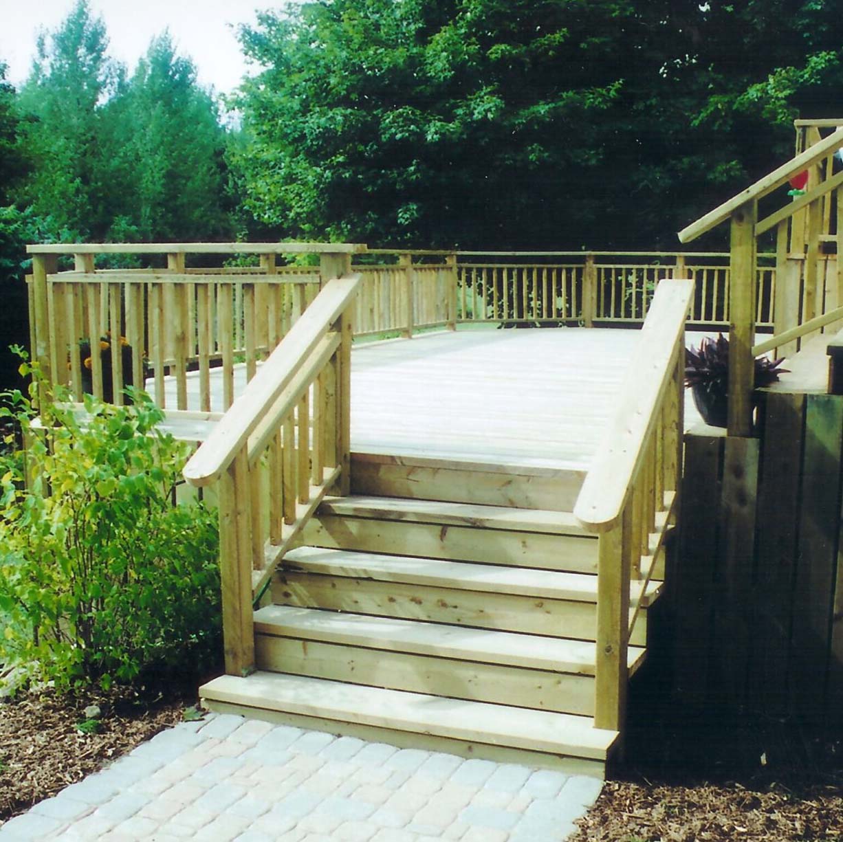 Wooden deck with stairs and railing, surrounded by greenery. Stone pathway leads to the deck, creating a natural outdoor setting. No people present.