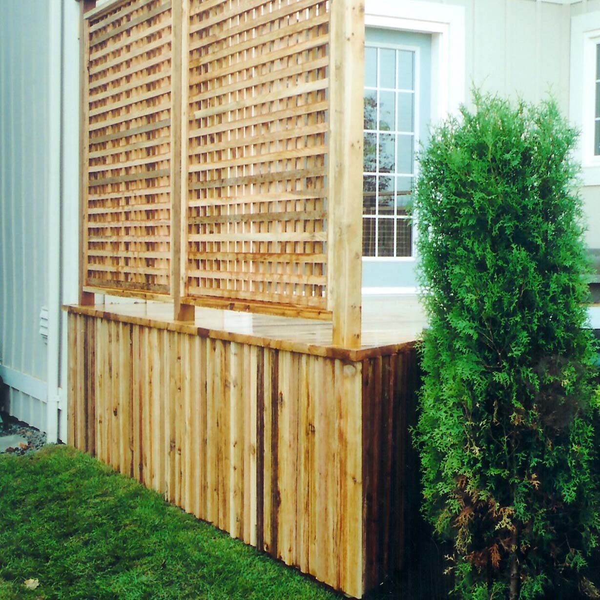 A wooden deck with lattice privacy screen and a lush green shrub, adjoining a house with blue door and windows. No people present.