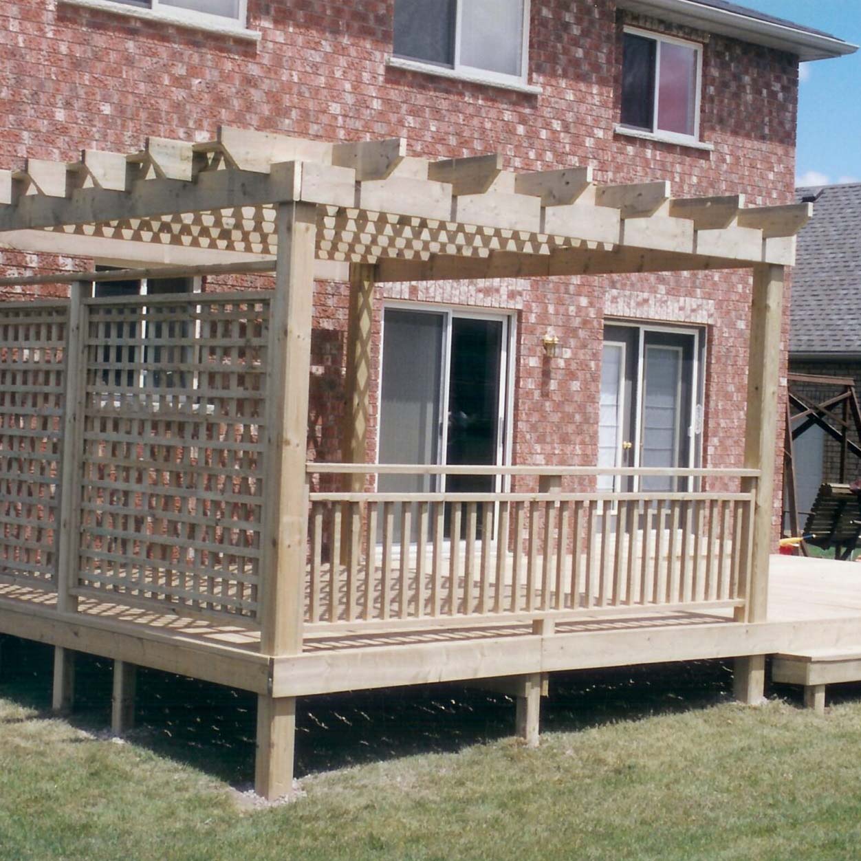 Wooden pergola on a raised deck, attached to a brick house with sliding glass doors. Sunny day, clear sky, grass below.