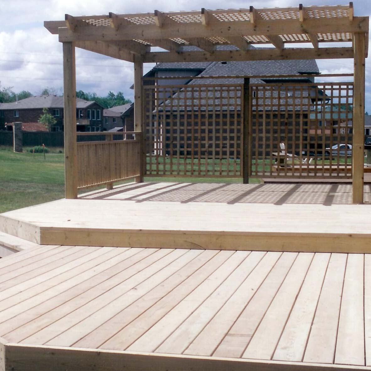 Wooden deck and pergola in a suburban backyard setting. Nearby houses and grassy area visible, under a partly cloudy sky. No landmarks identified.