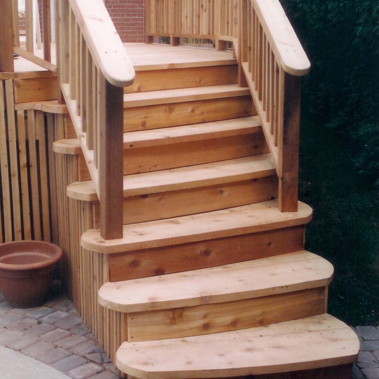 Wooden deck stairs with railings, leading from a patio. Potted plant on the side, brick wall in the background, surrounded by greenery.