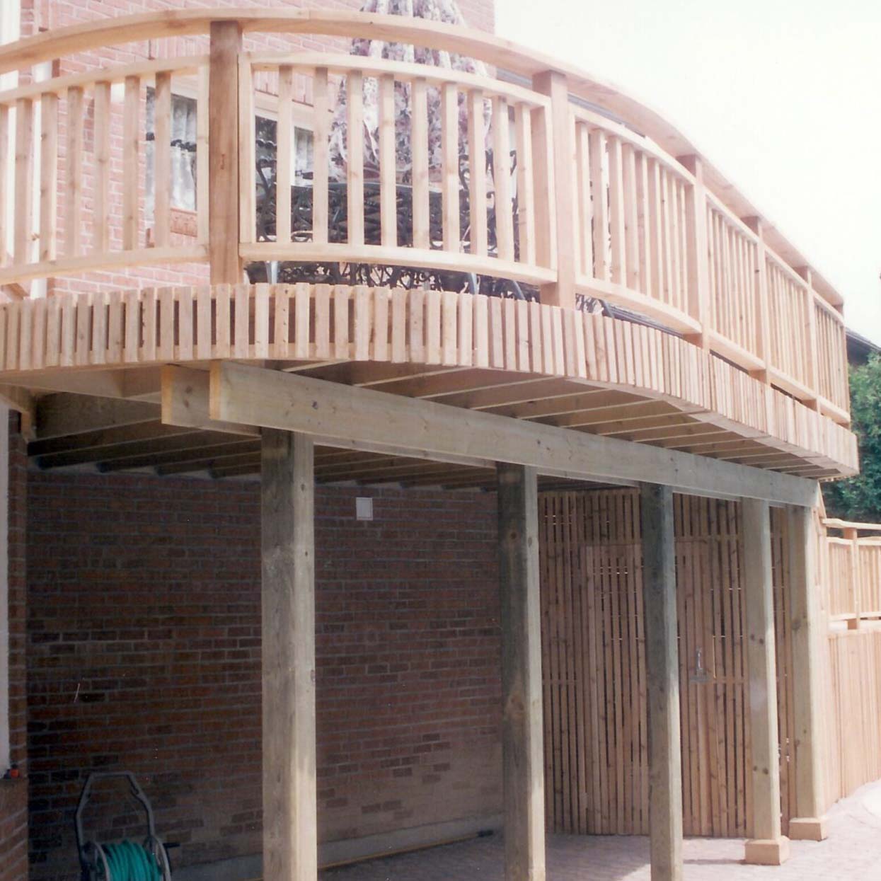 A wooden deck extends above a brick exterior, supported by sturdy beams. The setting is outdoors, with a hose coiled against the wall.