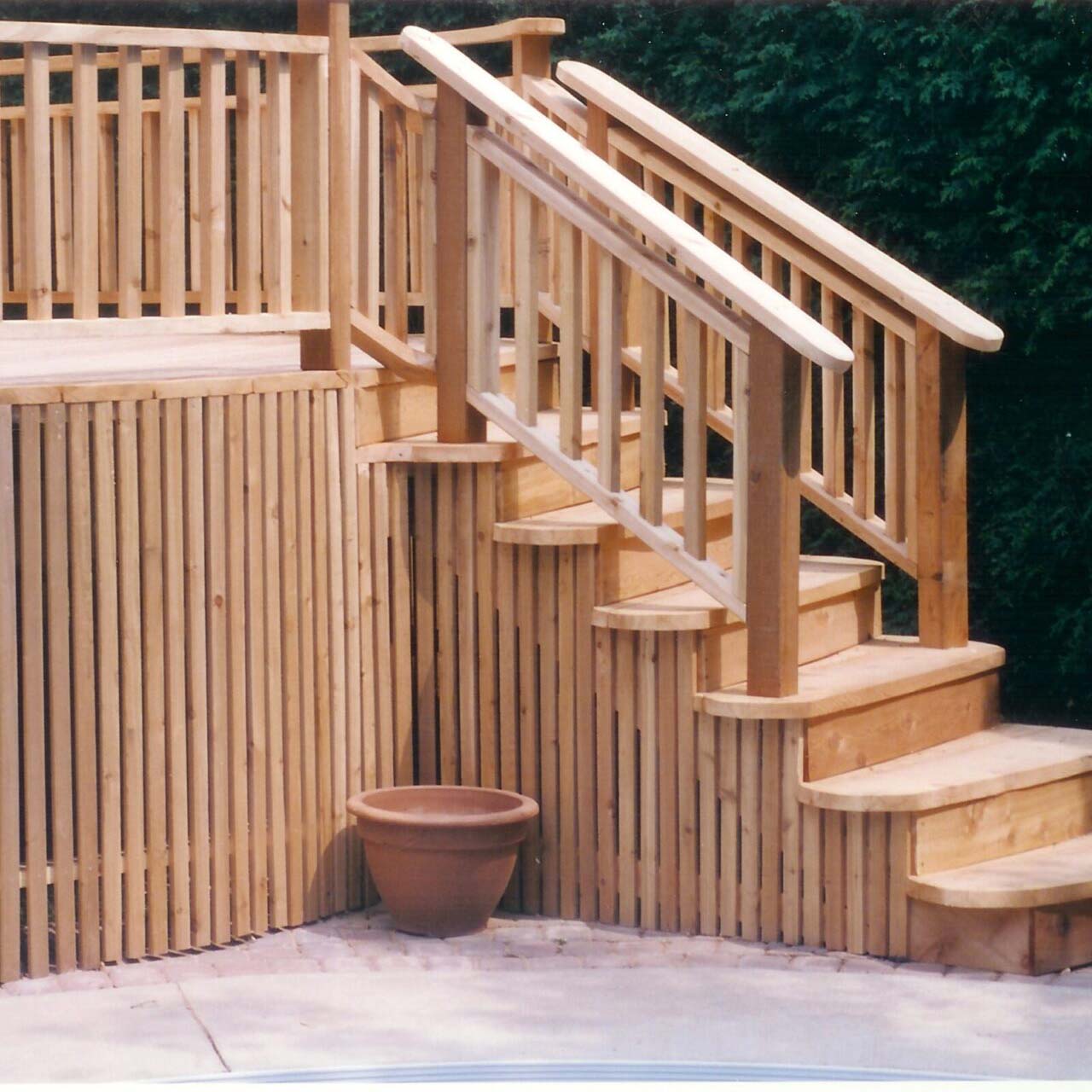 Wooden deck with railings and steps, accompanied by a terracotta planter. Surrounded by greenery, no people or recognizable landmarks are visible.